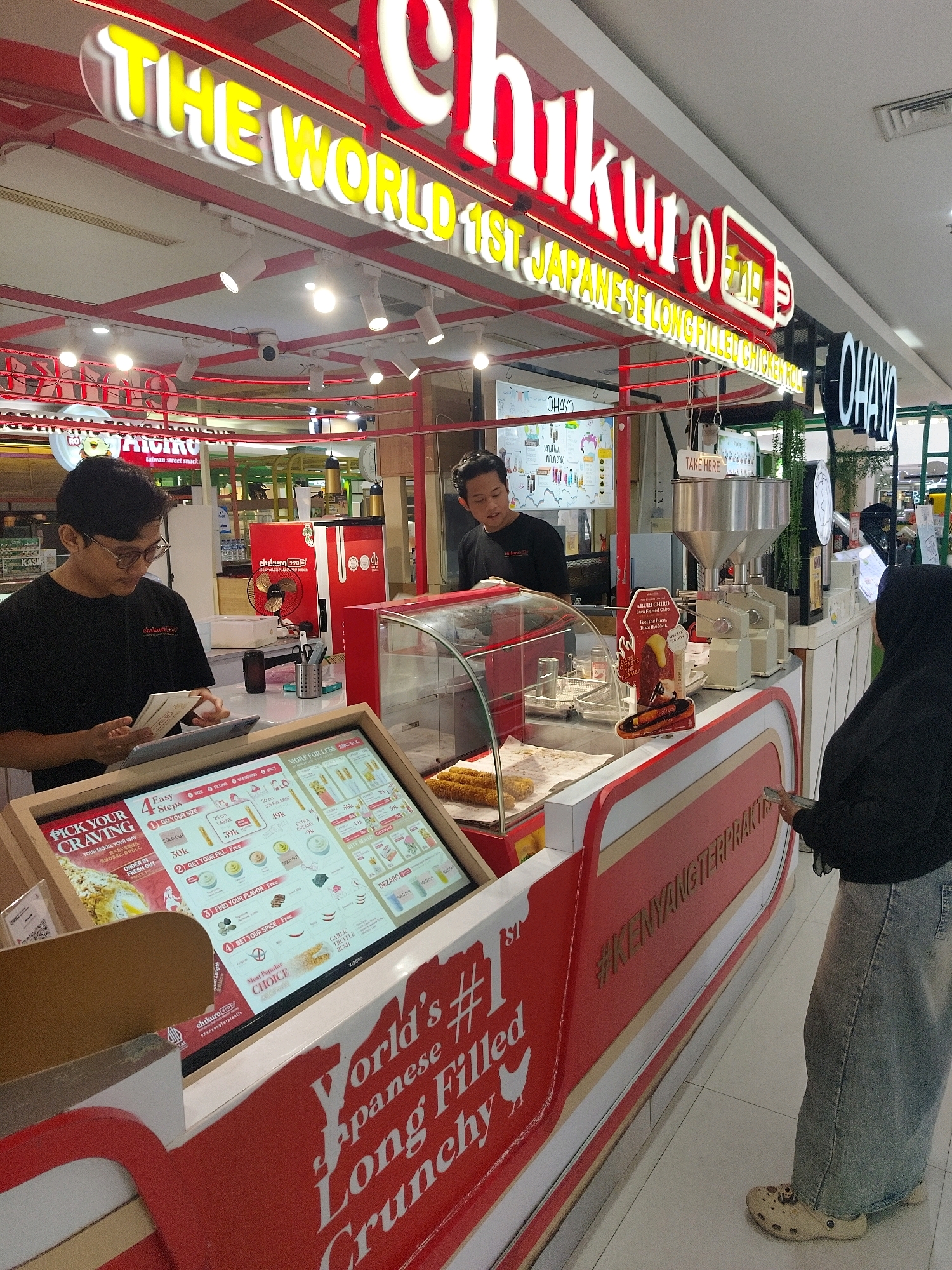 A wide shot shows a Chikuro food stall in a mall. The stall has a red and white striped exterior, a glass display case with food items, and a digital menu board. Two employees are behind the counter, one taking an order and the other preparing food. A customer stands in front of the counter, looking at the menu. The mall interior is visible in the background, with other stalls and people. A sign above the stall reads "THE WORLD'S 1ST JAPANESE LONG FILLED CHICKEN ROLL". The stall is located in the city of Mojokerto, Indonesia.