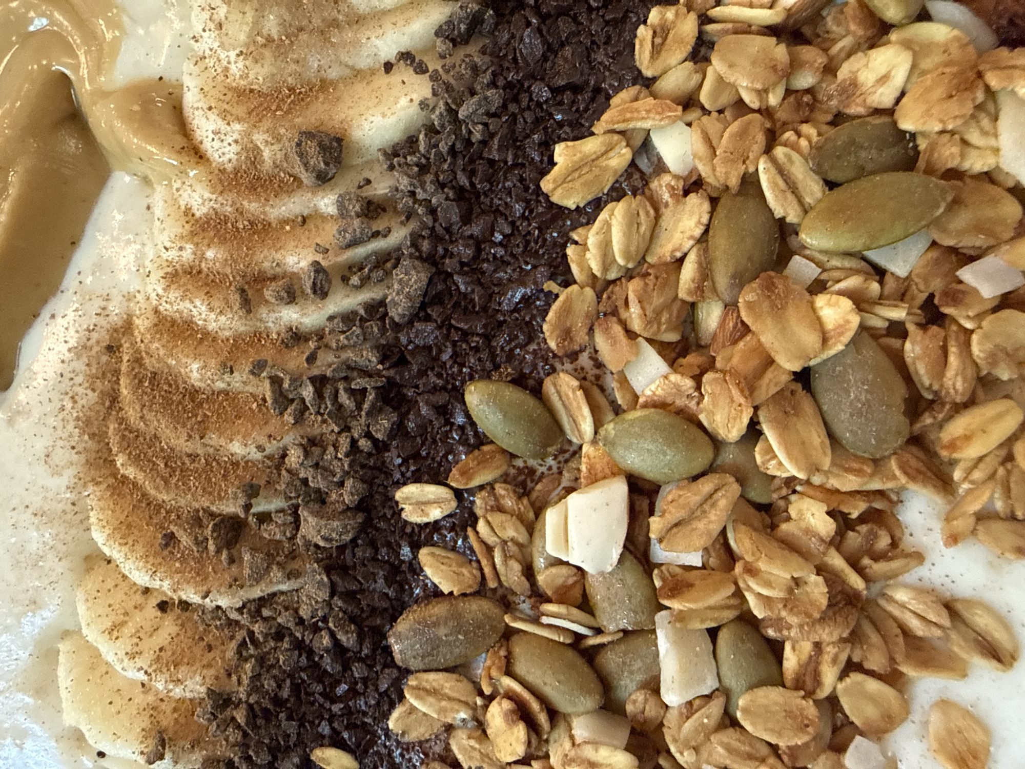 This is a close-up, overhead shot of a smoothie bowl. The bowl appears to be filled with a creamy, white base, likely yogurt or a smoothie. Layered on top are sliced bananas dusted with cinnamon, a dark, crumbled topping that looks like chocolate or coffee grounds, and granola with pumpkin seeds and white chunks, possibly dried coconut or white chocolate. A drizzle of a light brown, viscous liquid, resembling peanut butter or tahini, is visible at the top. There are no people, actions, or interactions visible. The environment is not depicted. The image does not provide any information about its location in Moscow, Russia.