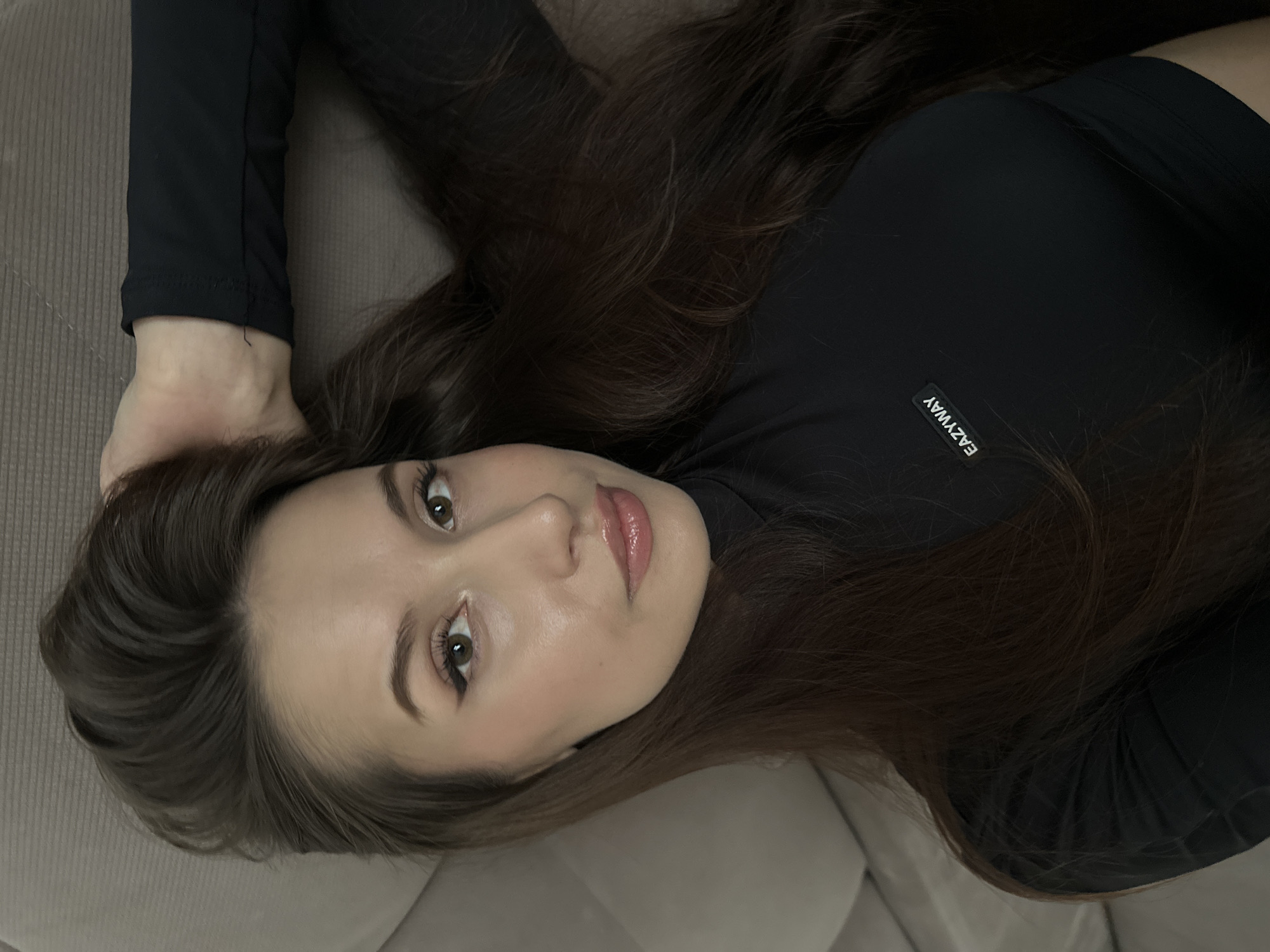 The image captures a close-up portrait of a young woman lying on her back on a light brown or beige upholstered surface, likely a sofa or bed. Her head is positioned towards the bottom left of the frame, and she gazes directly at the camera with a calm and direct expression.
She has long, flowing dark brown hair that spreads out around her head and over her shoulders, contrasting gently with her fair skin and the neutral background. She is wearing a black, long-sleeved top. A small, rectangular white label bearing the text "EAZYWAY" is visible on the upper chest or shoulder area of her top, on the viewer's left.
Her facial features include well-defined eyebrows, brown eyes enhanced with subtle eyeliner and mascara, and glossy, natural-toned pink lips. The lighting is soft and diffused, characteristic of an indoor setting, and casts no harsh shadows, contributing to a serene atmosphere. The activity depicted is a posed portrait. Although the image itself lacks specific geographical indicators, the provided context specifies the location as Yekaterinburg, Russia.