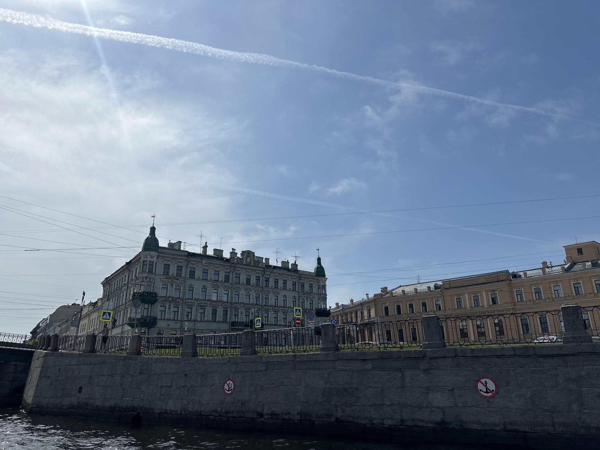 The image shows a view from a boat on a canal in Saint Petersburg, Russia. The foreground is dominated by the canal's gray stone embankment and the dark water.
Behind the embankment is a wrought-iron fence. Beyond the fence are two buildings. The one on the left is light-gray with several stories and decorative architectural details. It has green domes on its corners. A pedestrian crossing sign is visible in front of it. To the right is a light-yellow building, also multi-storied with architectural details. There appears to be a car parked near it.
The sky is blue with light clouds and contrails from airplanes. Power lines stretch across the upper part of the frame.