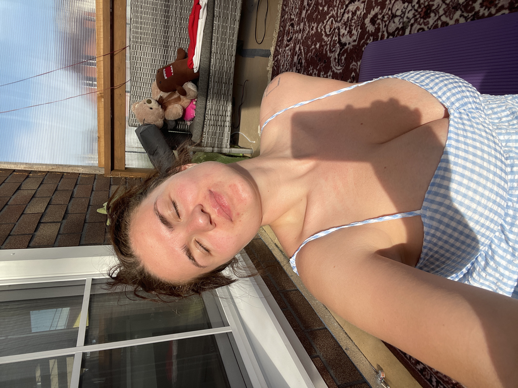 The image shows a woman lying on a purple yoga mat outdoors, in what appears to be a backyard or rooftop setting. She is wearing a blue and white checkered sundress. Her eyes are closed, suggesting she might be relaxing or sunbathing. Adjacent to the mat, there is a patterned rug and some outdoor furniture including what appears to be a rattan chair with stuffed animals on it. In the background, there are sections of a building with a window, and a clear, corrugated structure. A residential area appears in the distance. The exact location within the city of Мосметаллоконструкция Онт, Russia cannot be determined from the image alone.