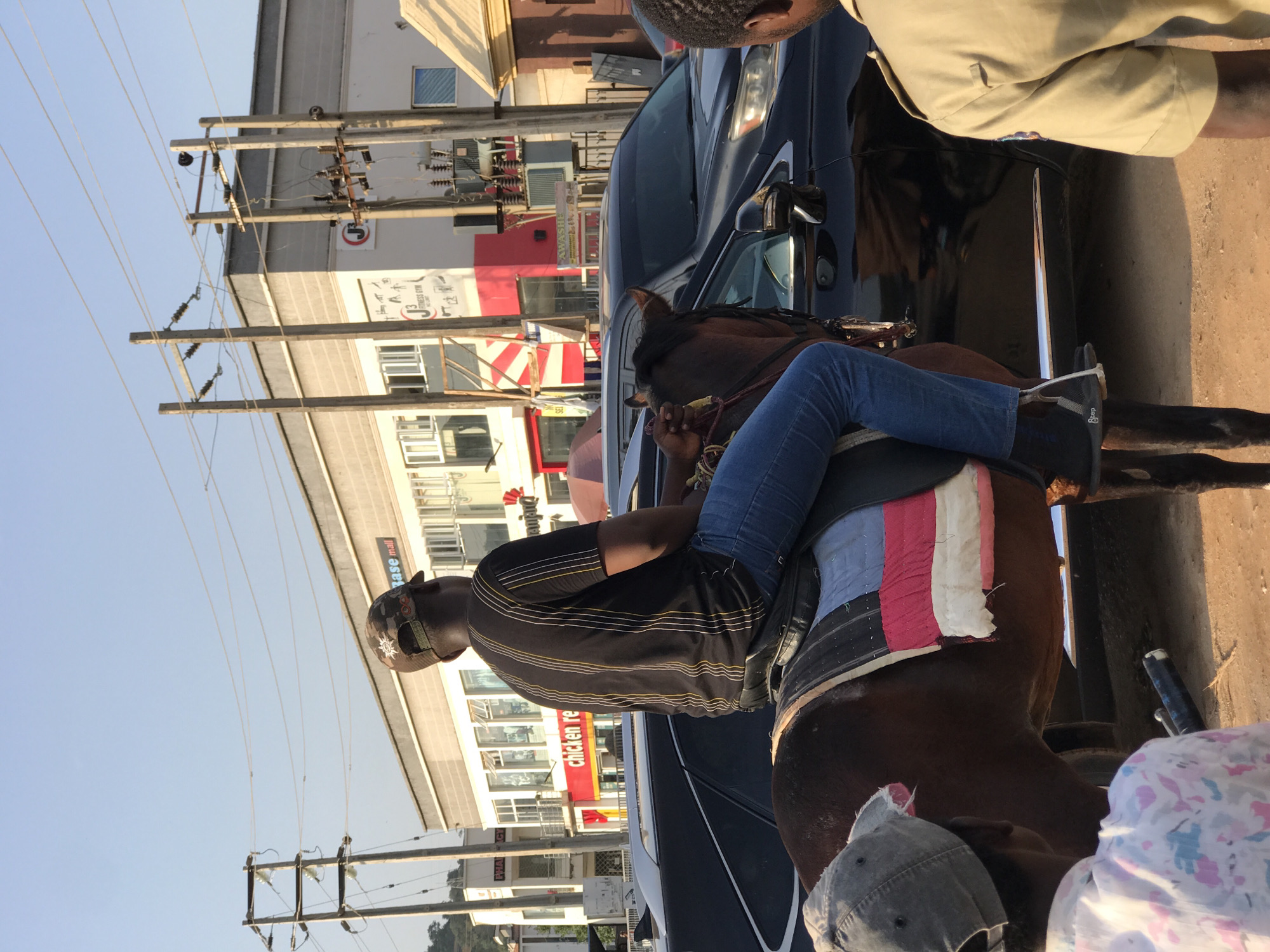 A man sits on a brown horse in Gwarinpa, Nigeria, amidst urban traffic. The man is wearing blue jeans, a dark striped shirt, and a baseball cap. He is seated on a saddle with a colorful pad. A dark-colored vehicle is parked next to the horse. In the background, there are utility poles with wires, buildings with commercial signs, including "Chicken Republic" and "Base Mall". Other people are nearby, partially visible in the frame. The weather appears sunny.