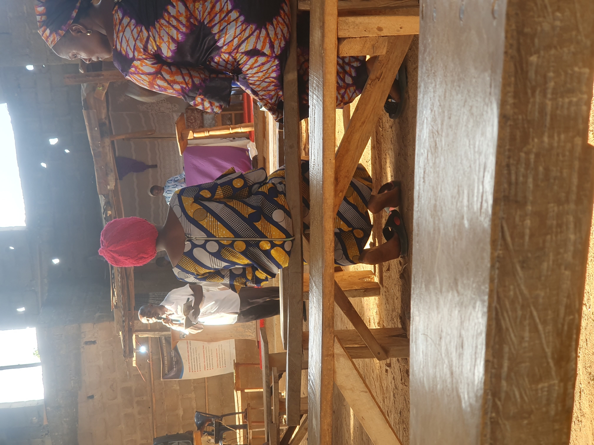 A sideways indoor shot shows a meeting or event held in what appears to be an unfinished building in Akwanga, Nigeria. Several people are present. Two women wearing colorful patterned clothing and head coverings are in the foreground. One wears a red head covering. A man in a white shirt speaks at a podium, where a sign titled "Catholic Biblical University Of Nigeria" is placed. The building's construction is visible with wooden beams, cinder block walls, and an incomplete roof. Sunlight streams in through openings. Chairs and other objects are visible in the background.