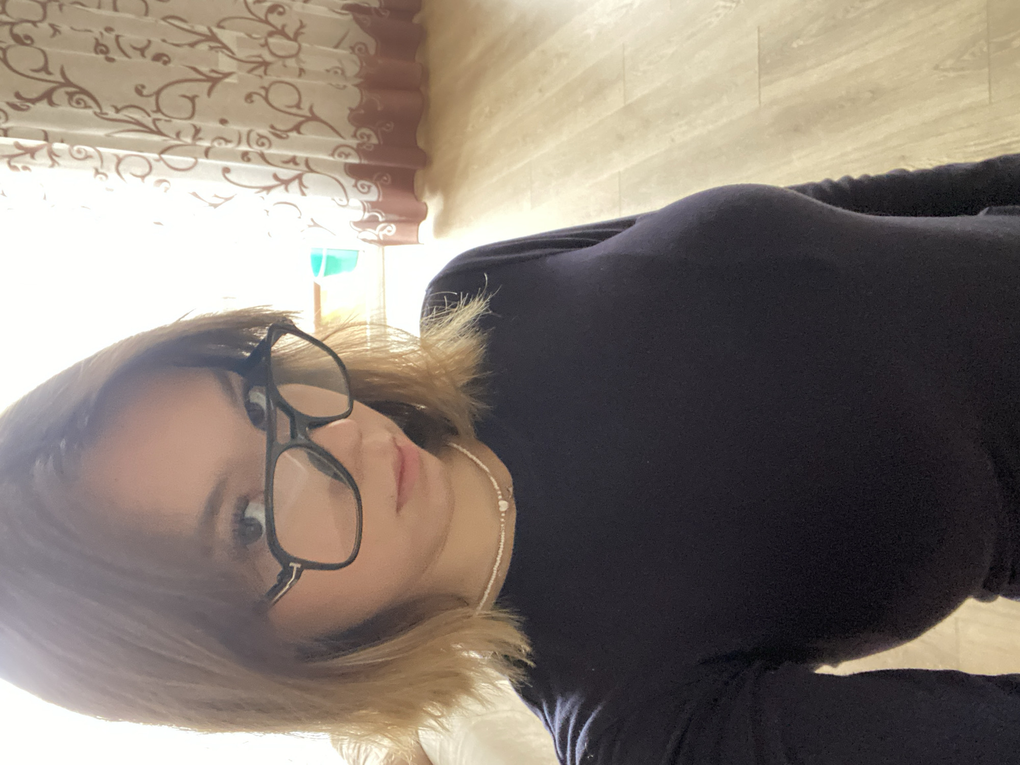 The scene is an indoor portrait featuring a young female, positioned centrally. She has shoulder-length, light brown hair with bangs and wears dark-framed eyeglasses with metallic accents on the temples. Her attire consists of a long-sleeved dark blue top and a light-colored beaded necklace with a small heart charm. She maintains direct eye contact with the camera. The background to her right shows a brightly illuminated window, partially obscured by a white curtain with a brown scroll pattern. A light blue bucket is partially visible on what appears to be a windowsill. The floor is constructed of light-toned wooden planks. A light-colored object is partially visible on the left. The specific location in the city of Chaikovsky, Russia, cannot be determined from the visual information provided.
