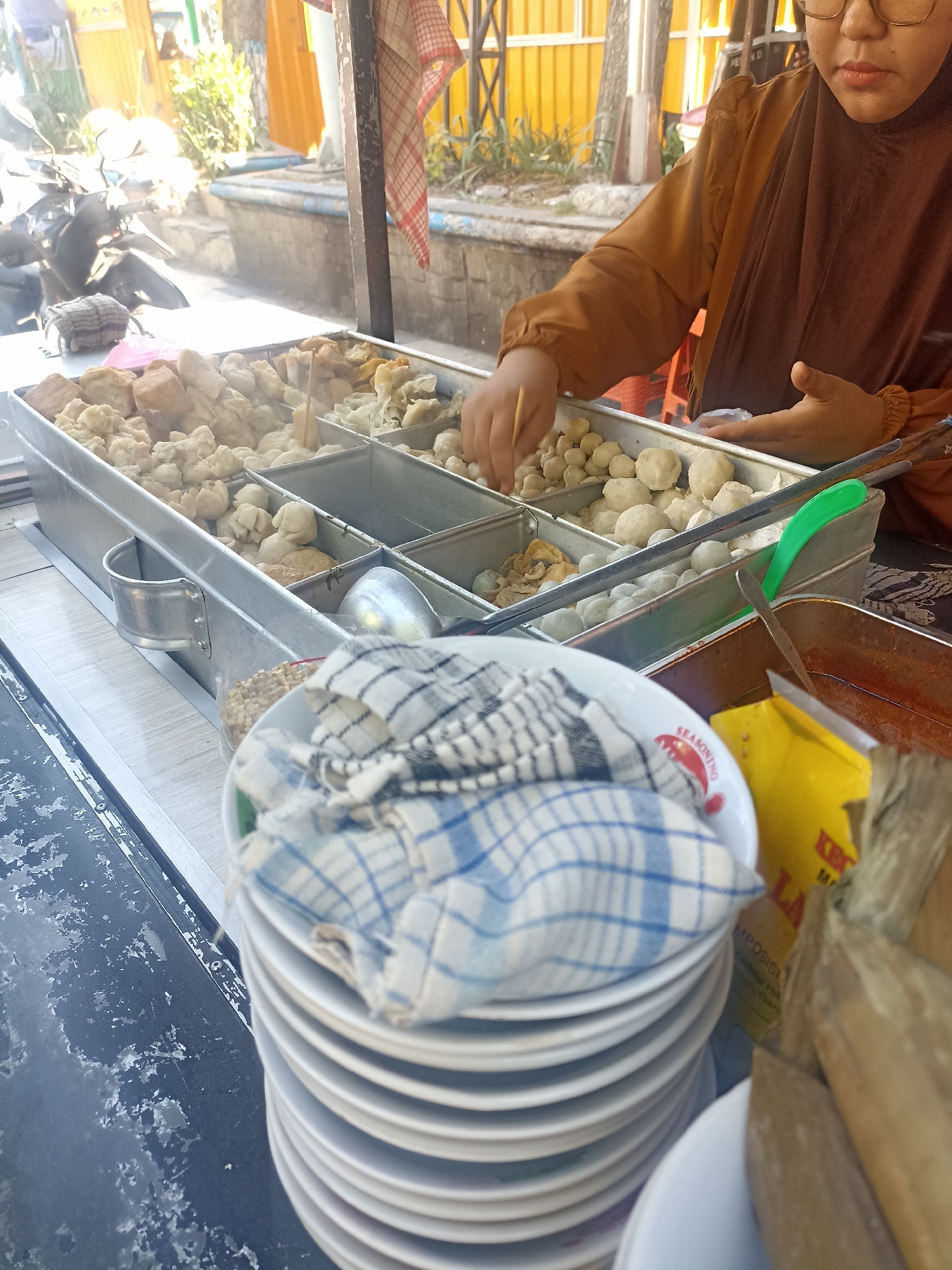 A street food vendor in Lamongan, Indonesia, is preparing food at a stall. The vendor, wearing a brown hijab and glasses, is seen from the chest up, reaching into a metal container filled with various food items. The stall displays multiple metal trays containing what appear to be meatballs and other fried or boiled food products. In the foreground, a stack of white plates is covered with a blue and white checkered cloth. Behind the food containers, a yellow package and a container of red sauce are visible. The background suggests an outdoor setting with a paved area and some greenery. The overall scene depicts a typical Indonesian street food vendor operation.