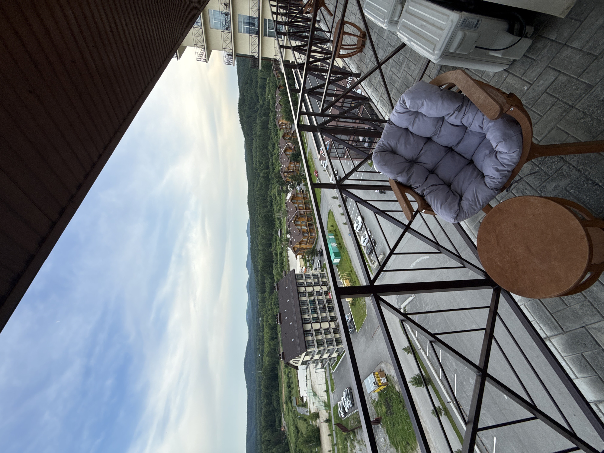 The image captures a panoramic view from a balcony overlooking a landscape. In the foreground, a wooden chair with a plush grey cushion is placed on a tiled balcony floor, next to a small wooden side table. Beyond the balcony railing, a multi-story apartment building with beige facade and balconies is visible, along with several smaller wooden houses with brown roofs. A paved road with parked cars runs in front of the buildings. The middle ground is characterized by a forest of green trees, and in the background, rolling hills under a cloudy sky complete the scene. The location is identified as Syrostan, Russia. There are no people visible in the image. The overall atmosphere is serene and picturesque, suggesting a peaceful retreat or residential area.