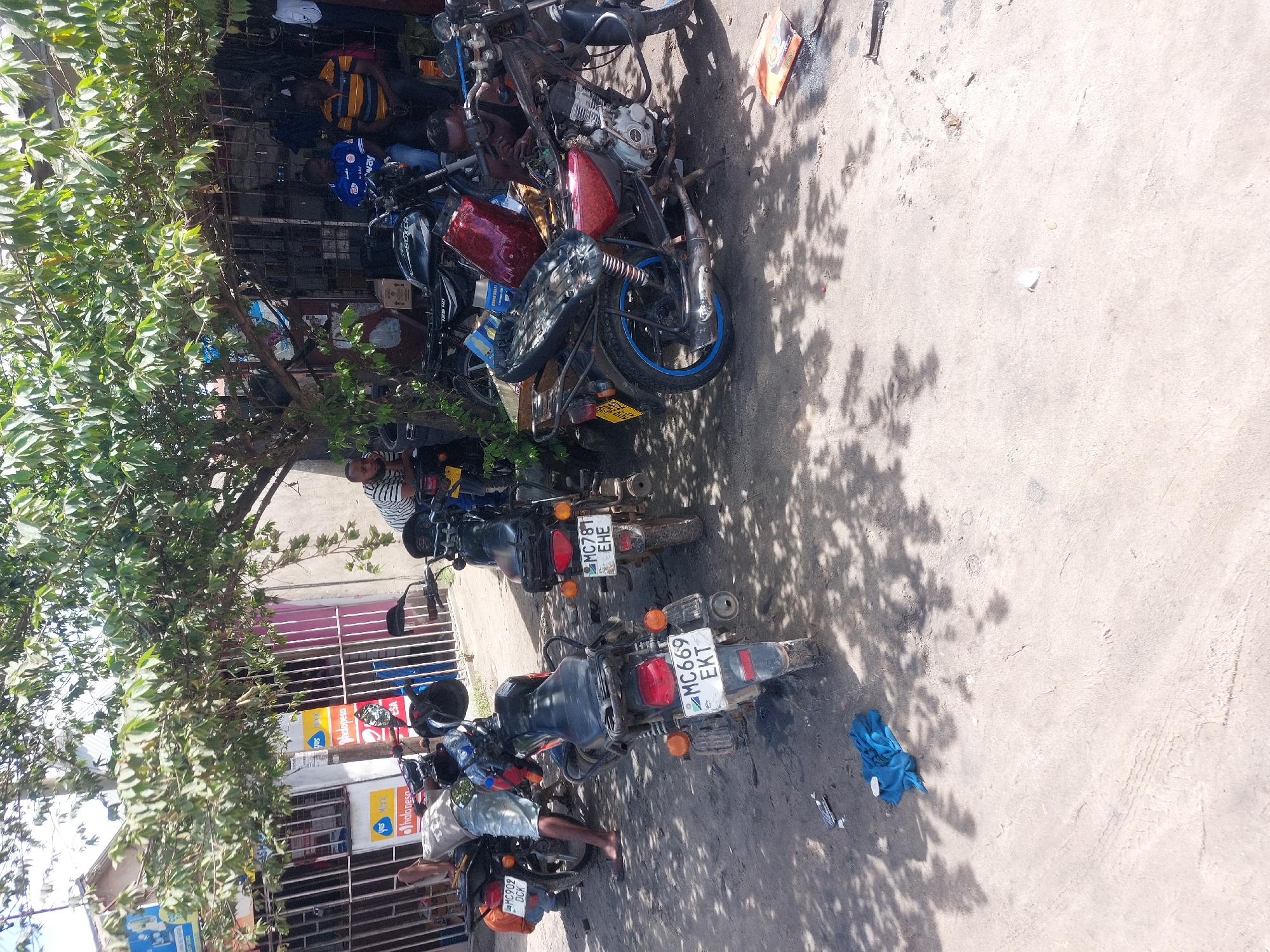 The image captures an outdoor scene in what appears to be a motorcycle repair shop or a street-side parking area in Dar es-Salaam, Tanzania. Several individuals are present among a line of motorcycles, some of which appear to be undergoing repair.
In the upper left, under the shade of a dense tree, a man in a yellow and black striped shirt and another in a blue shirt with "Away" visible are seated, possibly waiting or taking a break. Further down, another man in a striped shirt (white and black) leans against a parked motorcycle. Other figures are partially visible deeper within the shop's interior, suggesting activity.
The main subjects are numerous motorcycles, parked in a row along a concrete surface. Some are clearly in various states of repair, with parts exposed or removed. One prominent motorcycle in the foreground features a red fuel tank and a front wheel with a distinctive blue rim, appearing somewhat dismantled. Other motorcycles are black or dark-colored, with their rear ends facing the viewer.
The ground is concrete, marked with tire tracks and littered with a blue piece of cloth or garment near the center-right. Strong sunlight casts clear, defined shadows from the tree and the motorcycles, indicating a bright day, likely mid-morning or afternoon. The background reveals parts of buildings, possibly the repair shop itself, with corrugated metal grates and some commercial signage.
Visible text includes "Away" on a blue shirt and several motorcycle license plates: "MC609 DCK", "MC669 EKT", and "MC781 EHE". Partially visible signs in the background show "ndapesa" and "JOS", likely indicating mobile money services or a store. The scene depicts a bustling, everyday street or workshop environment.