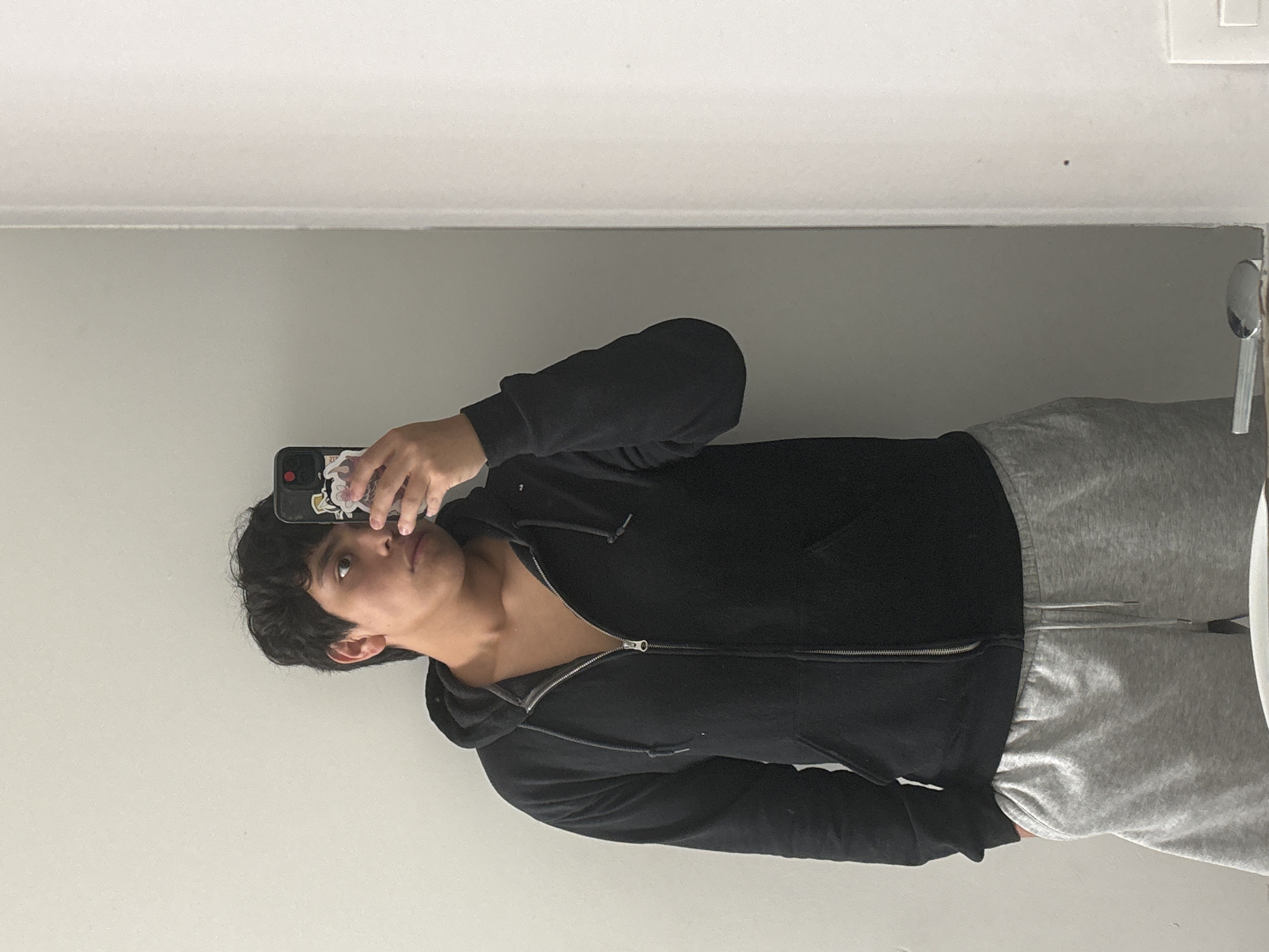 A young person is taking a selfie in a bathroom. The person has dark hair and is wearing a black zip-up hoodie and gray sweatpants. The background is a light gray wall and a white ceiling. A faucet and part of a mirror are visible on the right side of the frame. The person is holding a smartphone with stickers on the back. The location is unknown.