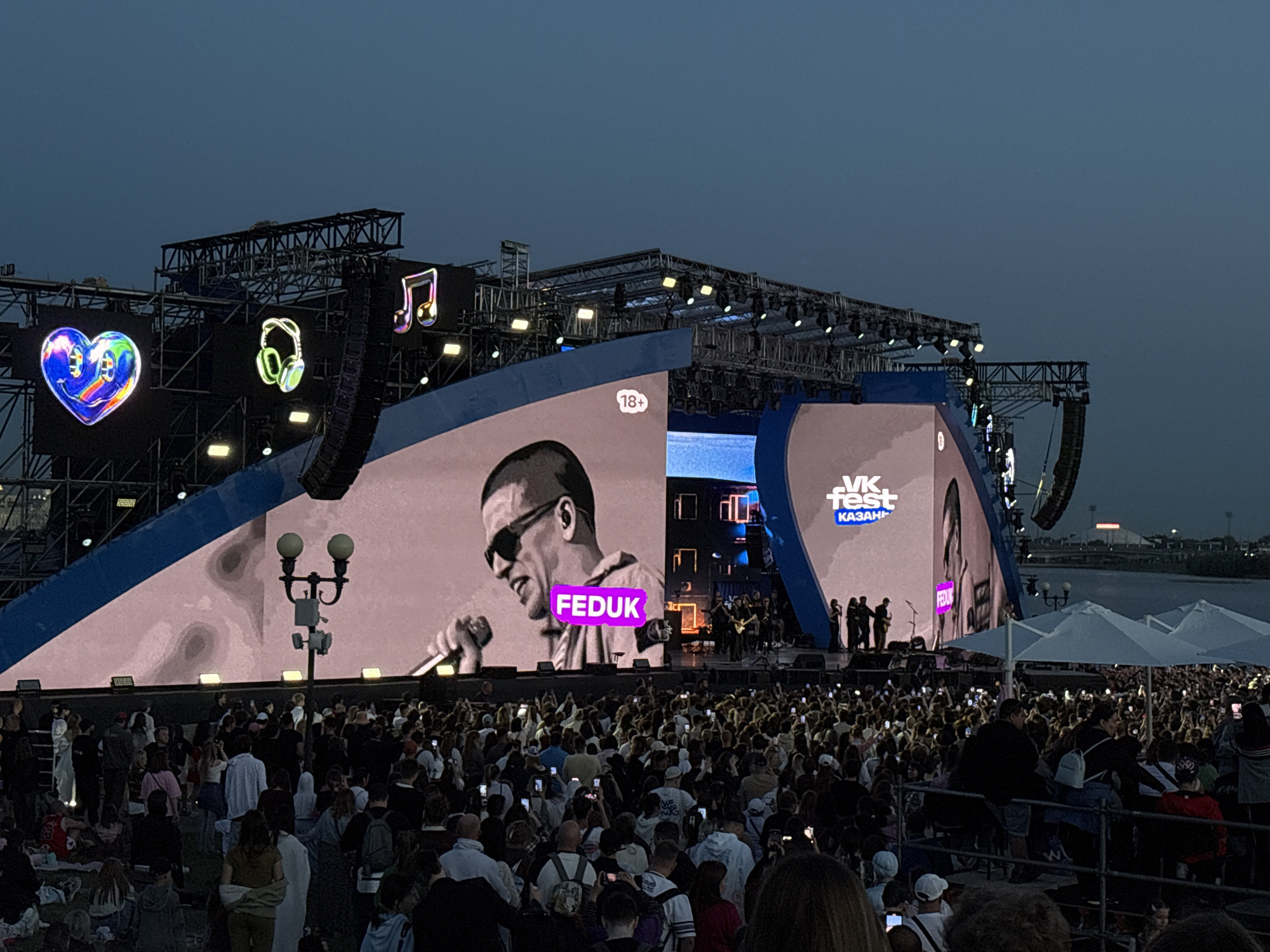 An outdoor music festival, identified as VK Fest Kazan, is depicted at dusk or early night in Kazan, Russia. A large, multi-panel LED stage dominates the scene, displaying a stylized, monochrome image of the Russian rapper Feduk, wearing sunglasses and holding a microphone, with his name "FEDUK" highlighted in a luminous pink box. An "18+" age restriction symbol is visible on the upper left screen panel. To the left of the main screen, above the stage, three illuminated holographic-style icons – a multicolored heart with a smiley face, green headphones, and a shimmering music note – are affixed to a metal truss structure.
A dense crowd of attendees occupies the foreground and midground, facing the stage. Numerous individuals are holding up smartphones, illuminated by their screens, indicating they are recording or photographing the performance. The crowd is diverse in appearance. To the right, behind the crowd, several white outdoor parasols are visible. The sky above is a dark blue-grey, signifying the time of day. In the background, beyond the stage, a body of water is discernible, with faint lights on the distant bank. The stage itself is constructed with extensive metal trussing, supporting multiple stage lights and large vertical speaker arrays on both sides. A black lamppost with two illuminated spherical globes stands in the midground, left of center, between the crowd and the stage.