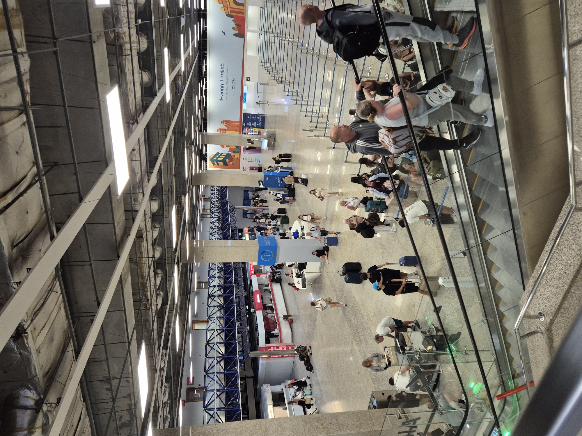The image depicts the interior of a modern airport terminal in Luqa, Malta. In the foreground, an escalator is visible with several people ascending it. One woman in the foreground is wearing a grey sleeveless top, grey pants, and white sneakers, carrying a backpack. A man behind her is wearing a grey hoodie and black pants. In the middle ground, numerous travelers are seen walking, standing, and sitting, some with luggage. Check-in counters and waiting areas with blue rope barriers are in the background. A large banner with "Il-vjaġġ it-tajjeb" and "Malta Airport" is displayed on the wall. The environment is well-lit with overhead fluorescent lights and appears to be a busy transportation hub.
