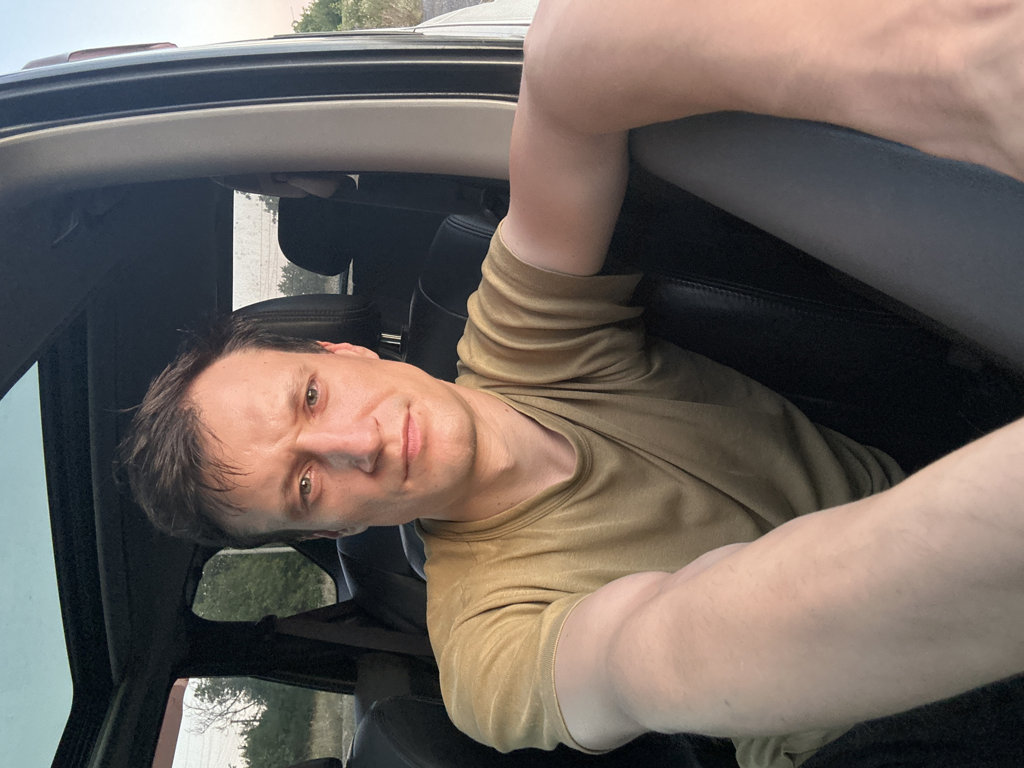 A medium shot, selfie-style photograph shows a white male with light skin and brown hair, looking directly at the camera. He is sitting in the back seat of a car with a sunroof open. He is wearing a tan t-shirt. The interior of the car is dark. Through the open sunroof and windows, trees and a light sky can be seen. The image was taken in Makiv, Ukraine, during the late afternoon or early evening, as indicated by the warm, golden light.