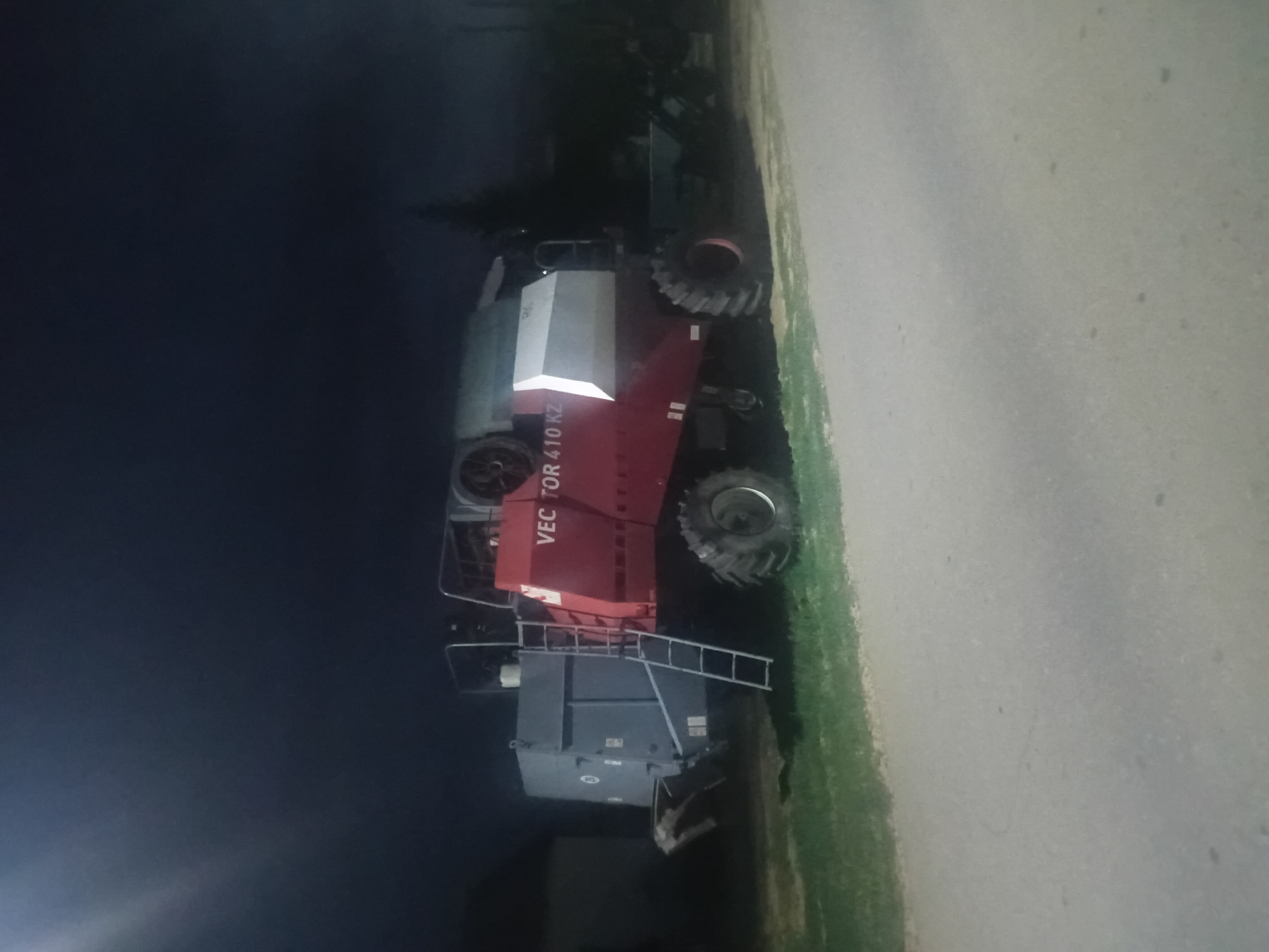 A red and white combine harvester, identified as a "VECTOR 410 KZ", is parked on the side of a road at night in Малая Владимировка ауылдық округі, Kazakhstan. The combine is situated on a patch of green grass next to a paved road. The background is dark, with a hint of a building to the left and trees and other farm equipment to the right. The sky is black, with a bright light source creating a glare in the upper left corner of the image. No people are visible in the scene.