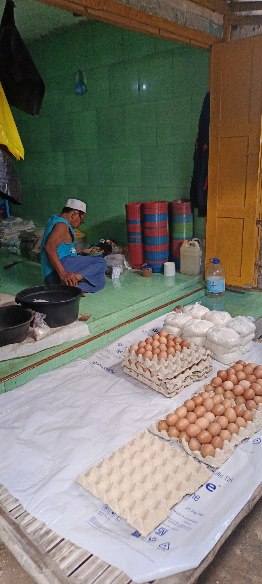 In the city of Gayam, Indonesia, a man wearing a white cap and a blue tank top sits on a green tiled floor. He is wearing blue pants and appears to be sorting through items. In front of him is a large black plastic tub. Behind him, stacked vertically, are several large cylindrical objects with alternating red and blue stripes. To his right, a yellow wooden door stands ajar, revealing a dark interior. The walls of the room are tiled in green. In the foreground, several egg cartons are stacked on white plastic sheeting. Some cartons are filled with brown eggs, while others are empty. Several bags of what appear to be rice are also visible behind the egg cartons. The scene suggests a marketplace or a storage area, possibly related to food sales or distribution.