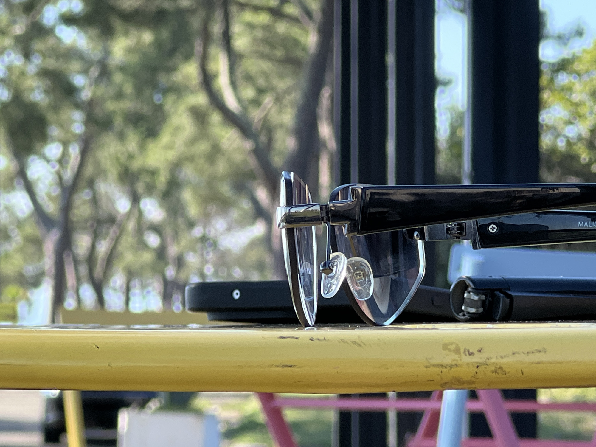 The image shows a pair of black-rimmed eyeglasses with clear lenses resting on a yellow surface. The eyeglasses are positioned on top of other dark objects, one of which appears to be a smartphone or similar electronic device. A black lighter with metallic accents is also visible next to the eyeglasses. The background is blurred, with hints of green trees and foliage, suggesting an outdoor setting. The lighting indicates it is daytime, possibly late morning or afternoon, with the sun casting shadows. The overall impression is a casual, still-life arrangement of personal items on what seems to be an outdoor table or railing. There is faint text visible on the arm of the eyeglasses, which reads "MALIS." The location context provided is Ochamchire, Abkhazia.