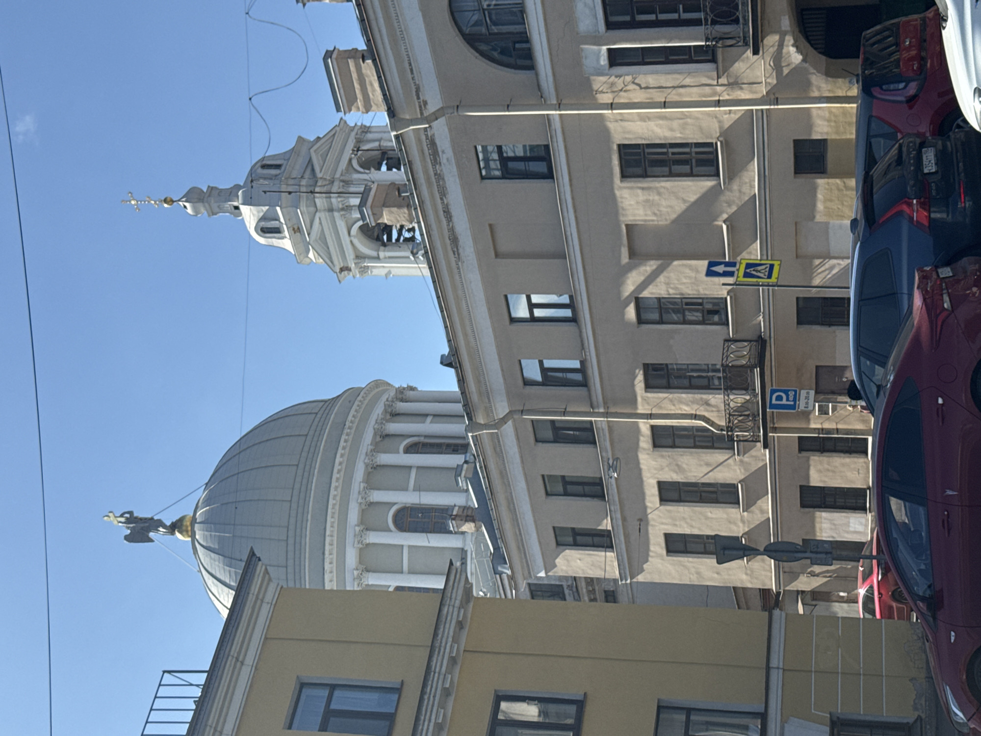 The image shows a street scene in Saint Petersburg, Russia. A large, domed building with white columns is visible in the background, with a spire rising behind it. The building is likely the Trinity Cathedral. In front of the cathedral, there is a beige building with several windows, and a parking sign is visible. Several cars are parked on the street, partially obscuring the building facades. The sky is blue.