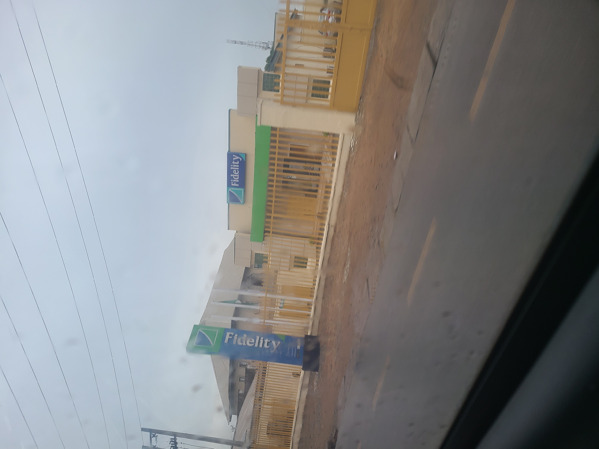 The image shows a Fidelity Bank branch in Karu, Nigeria. The bank building is painted in white and green, with a large yellow metal gate. A prominent blue and green Fidelity Bank sign is visible in front of the building. The road in front of the bank appears wet, suggesting it may have recently rained. The sky is overcast, indicating a cloudy day. The scene is likely during daytime, given the lighting. The image captures the exterior of the bank and its surroundings, which include a street and possibly other buildings in the background. There are no people clearly visible in the frame. The overall impression is that of a typical urban commercial setting in Nigeria.