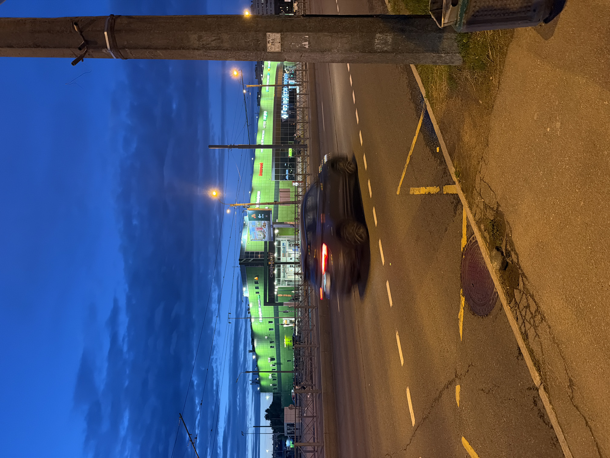 A motion-blurred dark colored SUV drives on a wet asphalt road at dusk. The road has white dashed lane markings and yellow lines painted on it. A concrete utility pole stands on the right side of the frame, with a metal trash can next to it. In the background, a large, brightly lit green building with signage is visible. The sky above is dark blue with clouds. The image appears to be taken from a sidewalk or elevated position next to the road.