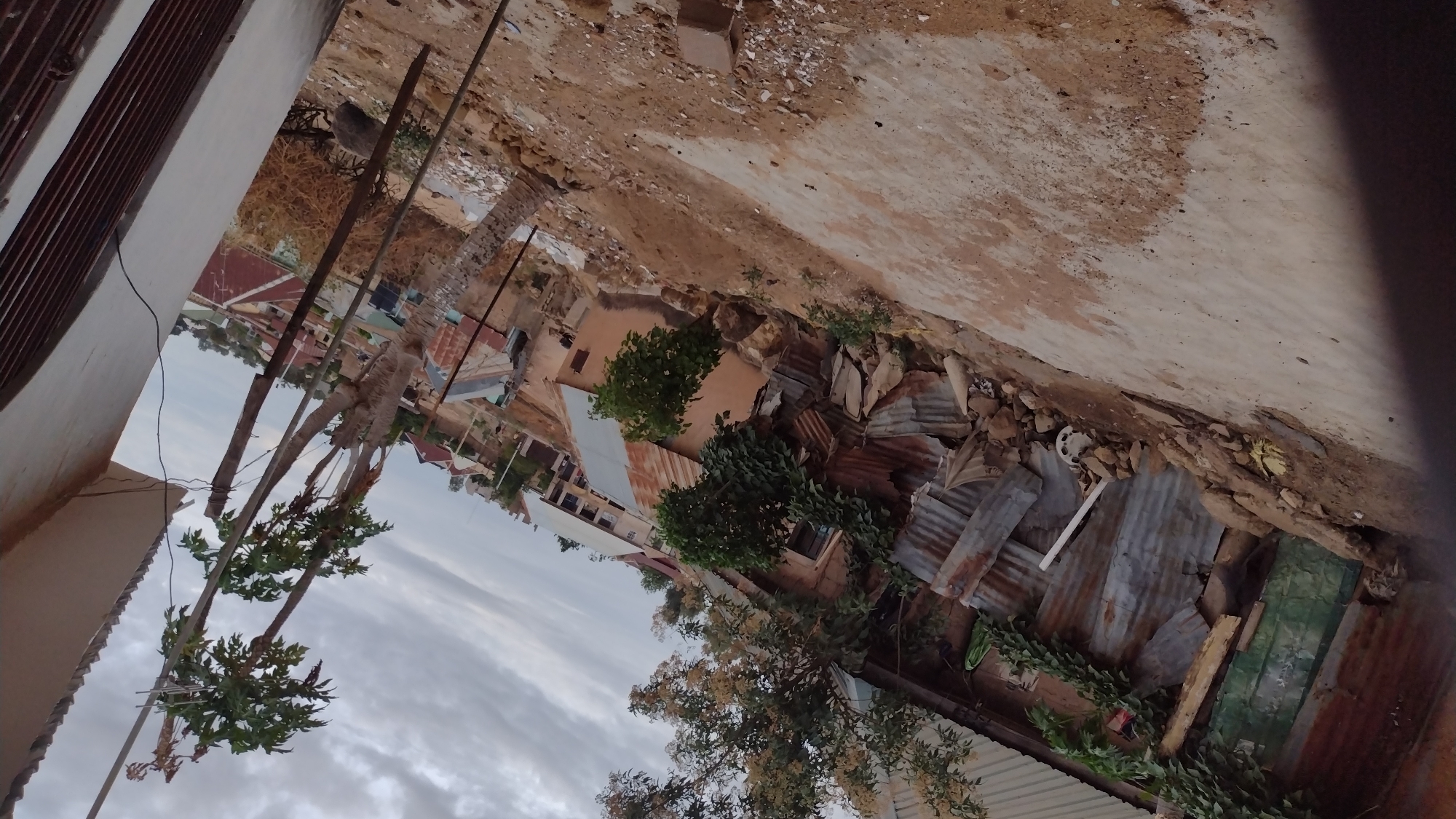 The image shows an elevated view of a densely built area, likely within the city of Jos, Nigeria. The foreground includes parts of a building and several papaya trees. The view then extends over corrugated metal rooftops intermixed with trees and vegetation. In the distance, more buildings and a dirt ground are visible. The sky is overcast with thick clouds.