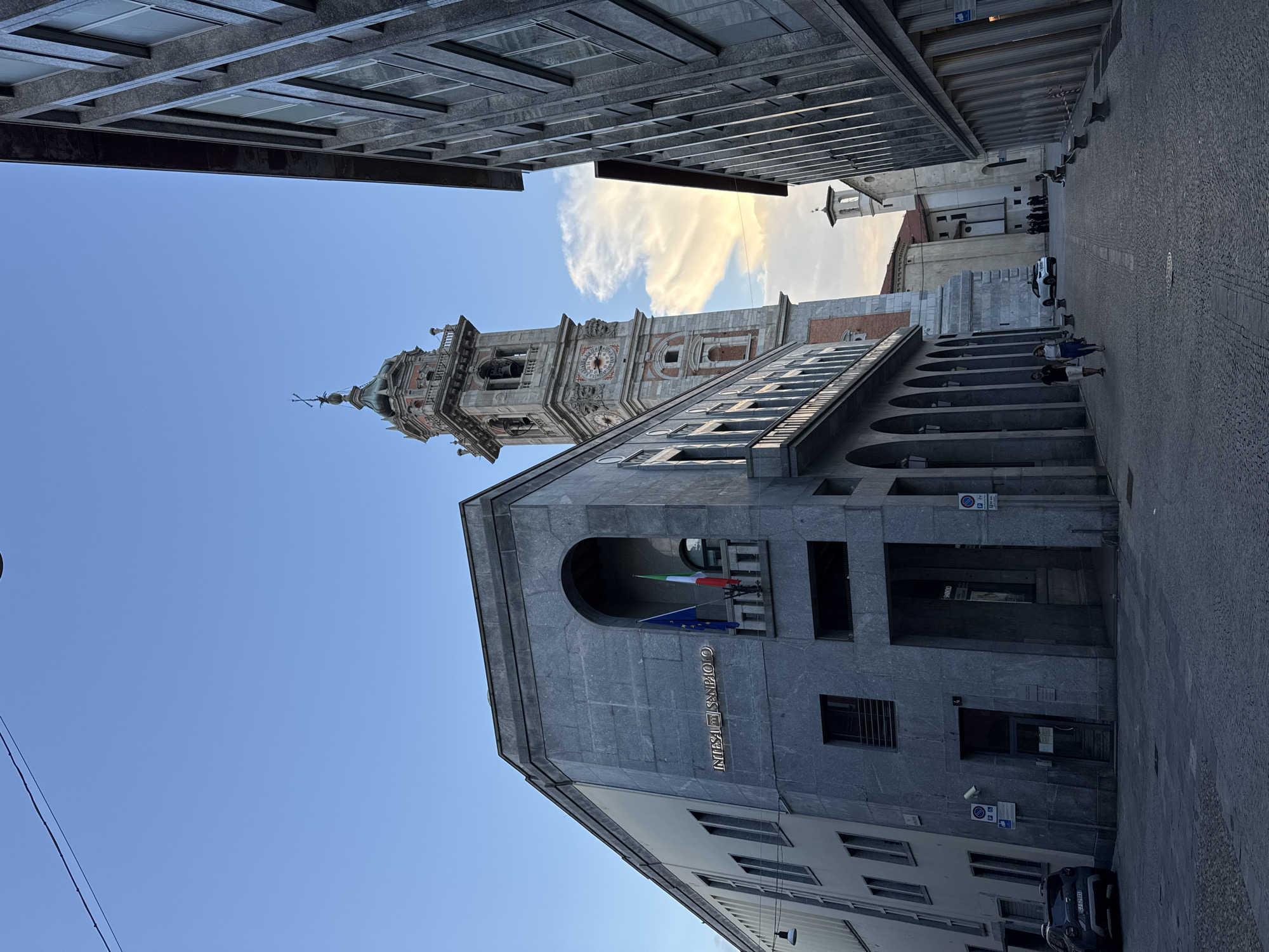 In Varese, Italy, the image depicts a street scene at dusk, dominated by the imposing architecture of the **Duomo di Varese** and the **Intesa Sanpaolo bank**. The Duomo's bell tower, a tall, ornate structure with a clock face, stands prominently in the background, catching the last rays of sunlight. The Intesa Sanpaolo building, a modern edifice clad in grey stone with arched windows and a distinctive overhang, occupies the left side of the frame, its signage clearly visible.
The street itself is paved with cobblestones, and on the right, another building with a facade of vertical grey panels lines the street. Two individuals, a man and a woman, are walking away from the camera on the cobblestone path. A few parked cars are visible, including a white SUV and a dark-colored hatchback. In the distance, other people can be seen congregating near a church entrance. The sky above is a clear blue, transitioning to warmer tones as it nears the horizon, indicating either sunrise or sunset. The overall atmosphere is one of urban tranquility as the day winds down.