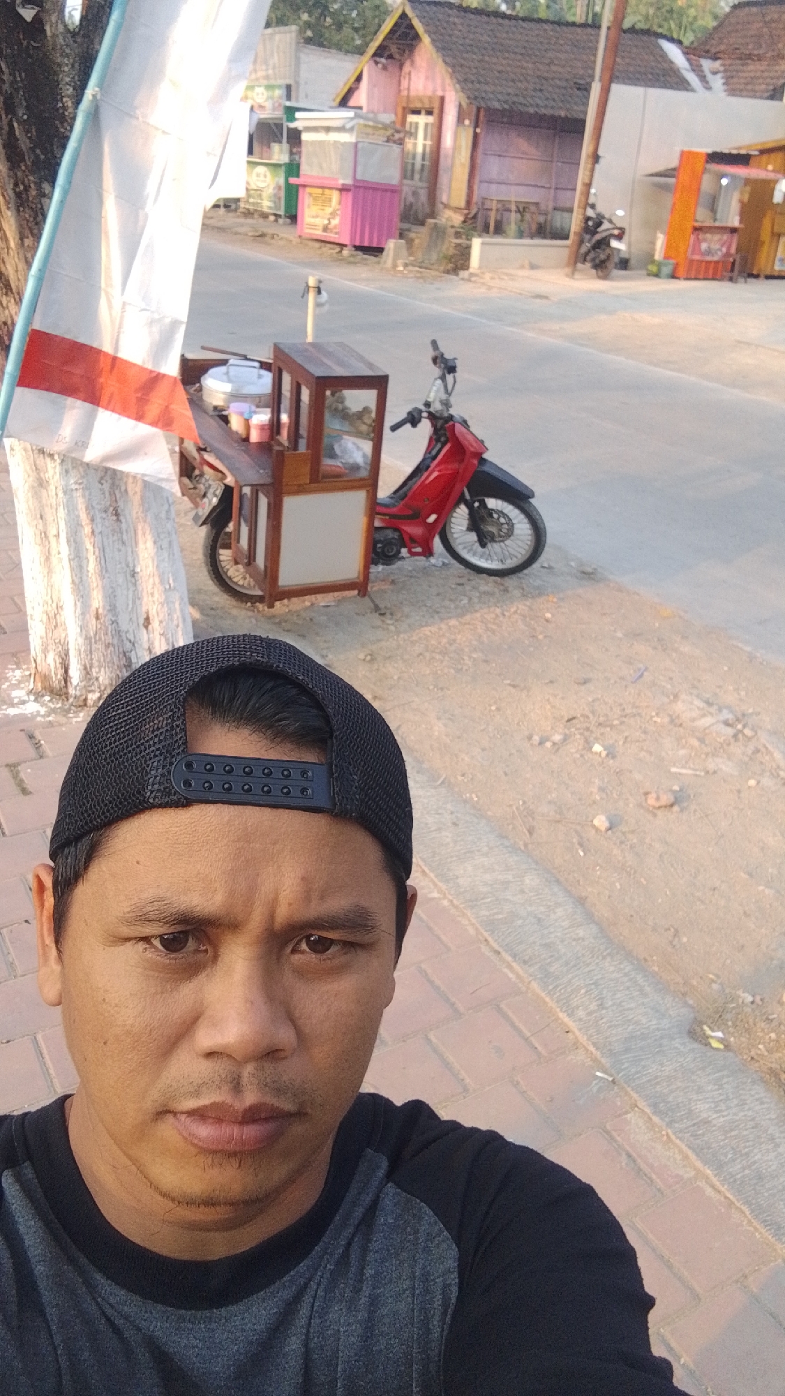 A man wearing a black baseball cap and a grey and black t-shirt takes a selfie outdoors in Karanglangu, Indonesia. The man is positioned in the foreground, with his face looking directly at the camera. Behind him, a red motorcycle is parked next to a small food cart. The food cart has a wooden frame and glass panels, and appears to be stocked with various food items. In the background, there are several small buildings, some of which seem to be shops or stalls. The ground is a mix of dirt and paved areas. The scene appears to be a street or roadside in a semi-rural or suburban area, with the warm light of what appears to be late afternoon or early morning casting long shadows.