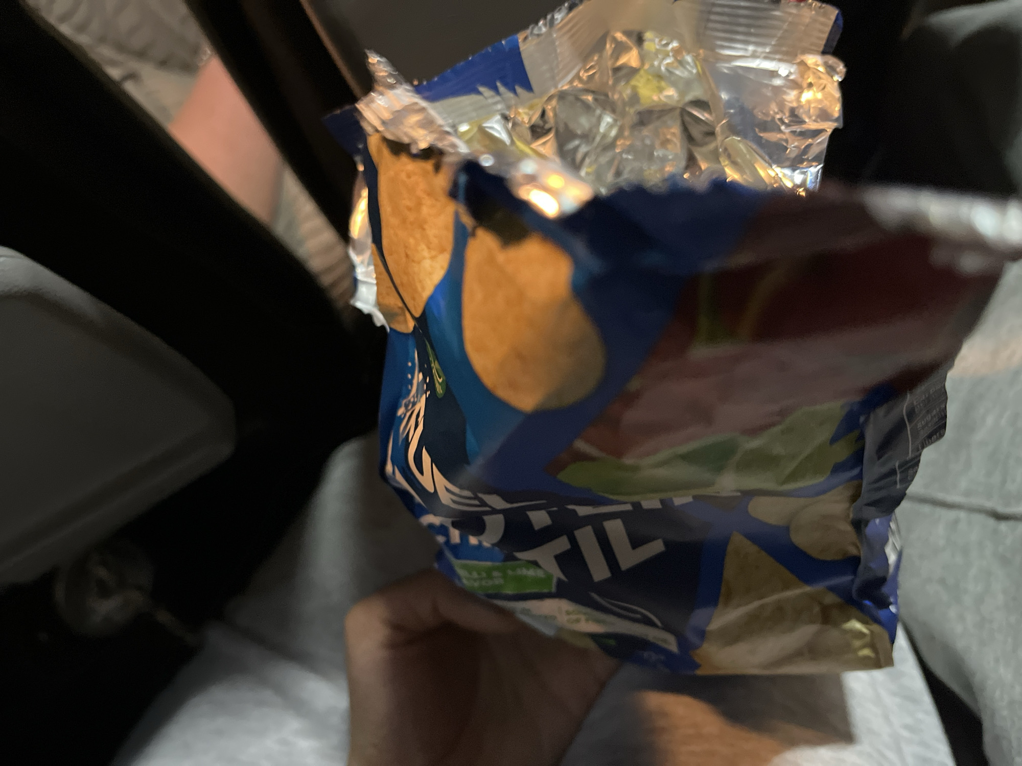 A partially opened bag of tortilla chips is being held in a person's hand inside a car. The bag is blue with a silver, crinkled top revealing the chips inside. The car's interior is visible in the background, including the seat, console, and door. The location cannot be determined from the provided image.