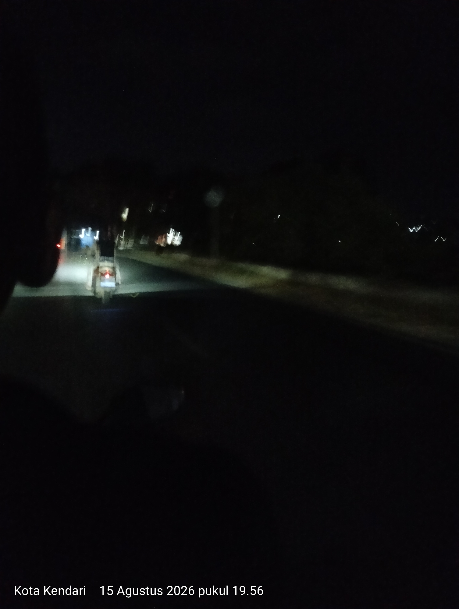 The image, captured at night, shows a blurry, dark scene on a road in Kendari, Indonesia. A motorbike is visible in the center of the frame, with its headlight illuminating the road ahead. The motorbike appears to have a rider, though their features are indistinct due to the low light and motion blur. The road surface is visible, and there are other blurred lights and shapes in the background, suggesting the presence of other vehicles or buildings. The text overlay at the bottom of the image reads "Kota Kendari | 15 Agustus 2026 pukul 19.56", indicating the location and the timestamp of the photograph. The overall impression is a snapshot of nighttime travel on a city road.