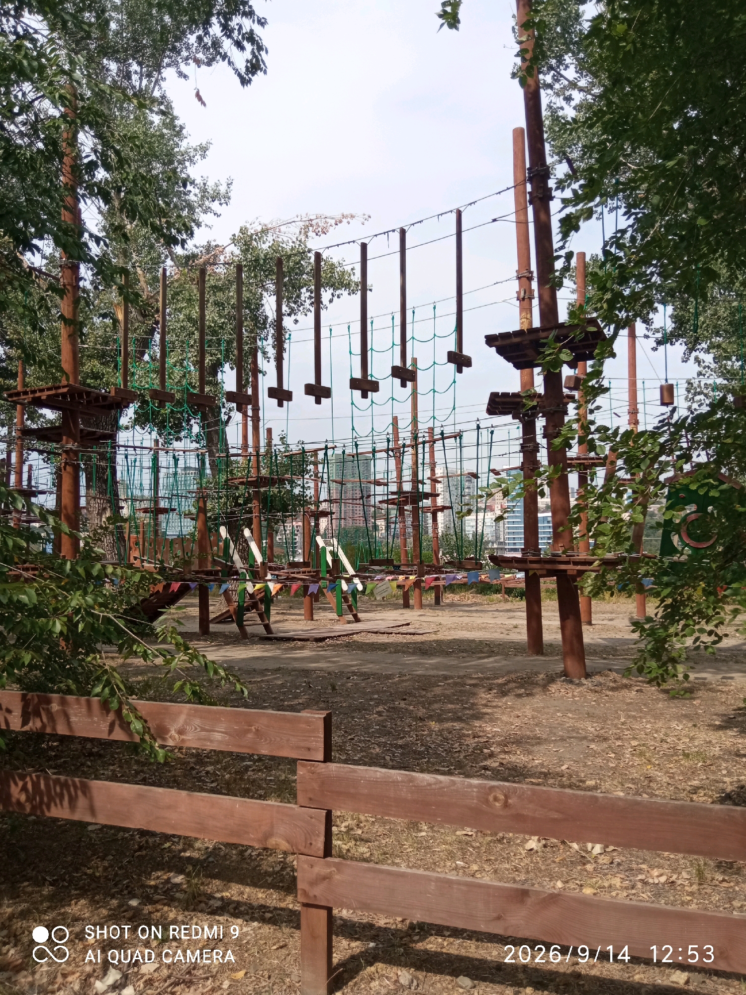 The image shows an outdoor adventure park, likely a rope course, with wooden platforms, ropes, and nets suspended between tall wooden poles. Several rope bridges and obstacles are visible. In the background, there are trees, and beyond them, modern multi-story buildings can be seen, suggesting an urban setting. The ground is covered with dirt and fallen leaves. In the foreground, a wooden fence runs horizontally across the bottom of the frame. A timestamp and camera information are overlaid on the lower part of the image. This scene is consistent with an outdoor recreational area located within the city of Rostov-on-Don, Russia, possibly near a waterfront or park area, given the presence of urban architecture in the background. There are no people visible in the image.