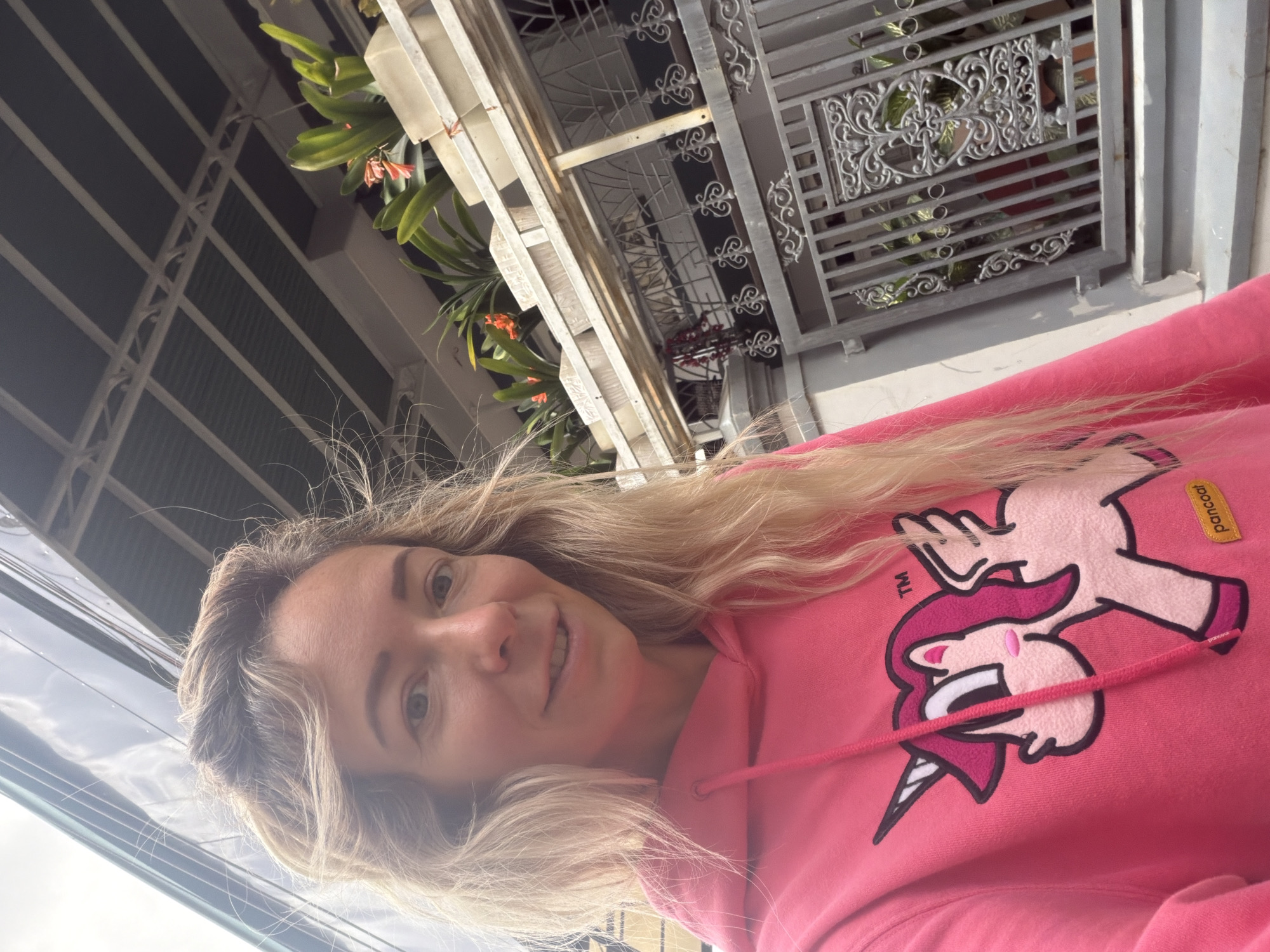 A young Caucasian woman with blonde, wavy hair is captured in a close-up selfie. She is wearing a bright pink hooded sweatshirt with a cartoon unicorn graphic on the front and a small yellow "pancoat" label. The woman is smiling and looking directly at the camera. The background suggests an outdoor setting, likely a residential area, with a building featuring a corrugated metal roof, a metal railing with decorative scrollwork, and several potted plants with orange flowers. The architecture and greenery are consistent with the style found in Da Lat, Vietnam. The lighting indicates it is daytime, with soft shadows.