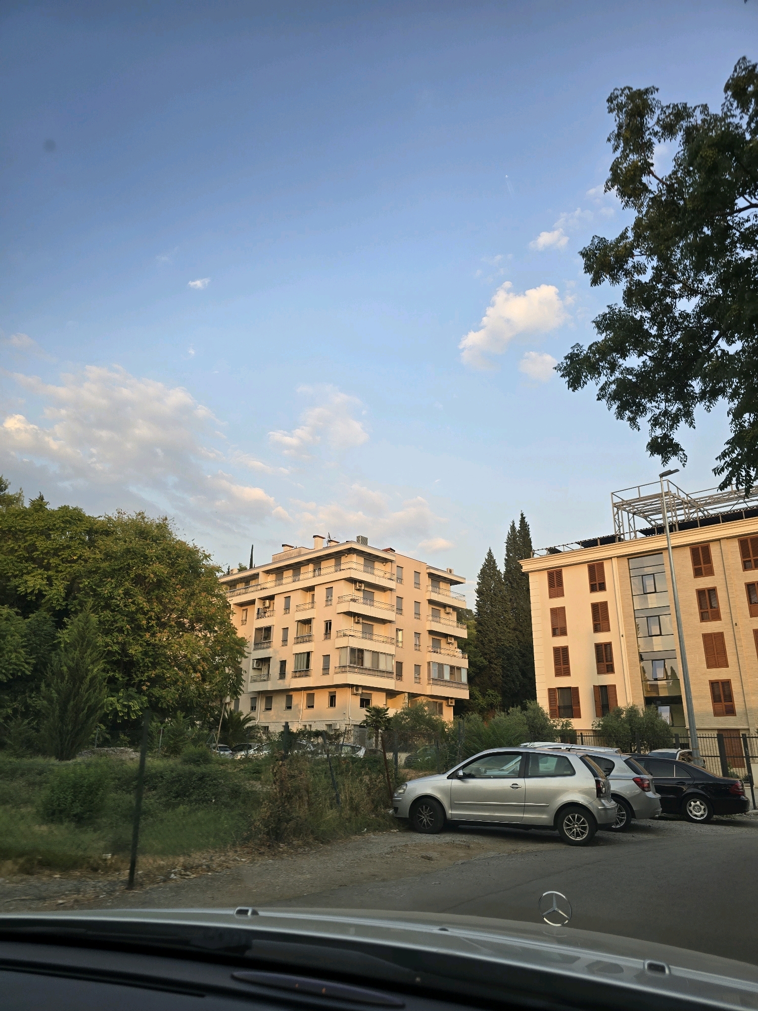 The image depicts a view of apartment buildings in Podgorica, Montenegro, with cars parked in front. The sky is clear with some scattered clouds, and the sun is low in the sky, casting a warm light on the buildings. The foreground shows the interior of a car, looking out at the scene. The buildings appear to be modern residential structures, with balconies and numerous windows. Trees and greenery surround the buildings, suggesting a suburban or semi-urban setting. The parked cars include a silver hatchback, a silver sedan, and a black sedan. The overall impression is one of a peaceful, late afternoon in a residential area of Podgorica.