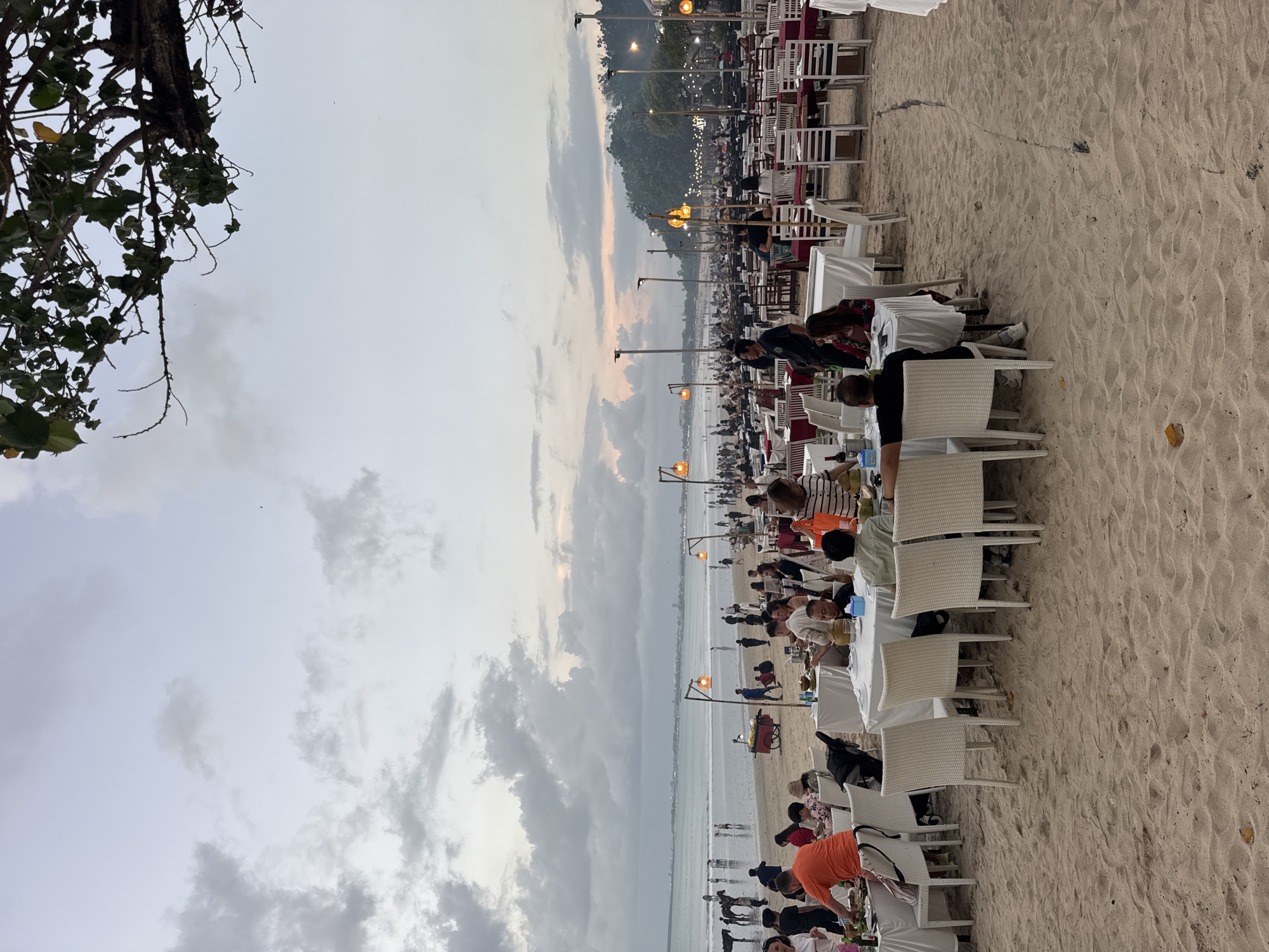 This image depicts a vibrant outdoor dining scene on a sandy beach during dusk or early evening in Kuta Selatan, Indonesia. Rows of white tables, predominantly covered with white tablecloths and paired with white chairs, are set up directly on the sand, extending from the foreground towards the water. Some tables also feature red accents, possibly tablecloths or runners.
Numerous people are seated at these tables, engaged in dining and socializing, creating a lively and bustling atmosphere. Other individuals are visible walking along the shoreline or moving between the dining areas. The patrons are casually dressed, fitting the relaxed beach environment.
The scene is illuminated by several tall poles topped with glowing orange-yellow lantern-style lights, which provide artificial light as the natural light of the day fades. The sky above is overcast with clouds displaying gradients of grey, light blue, and subtle orange or pink hues near the horizon, indicative of twilight or a recent sunset. In the background, the calm ocean stretches out, and a dark landmass or distant hills are visible across the water. Dark green foliage from an overhanging tree partially frames the image on the left. The sandy ground shows the textured patterns of footprints and ripples, common on a beach. A few small vendor carts can also be spotted closer to the water's edge. No discernible text is visible in the image.