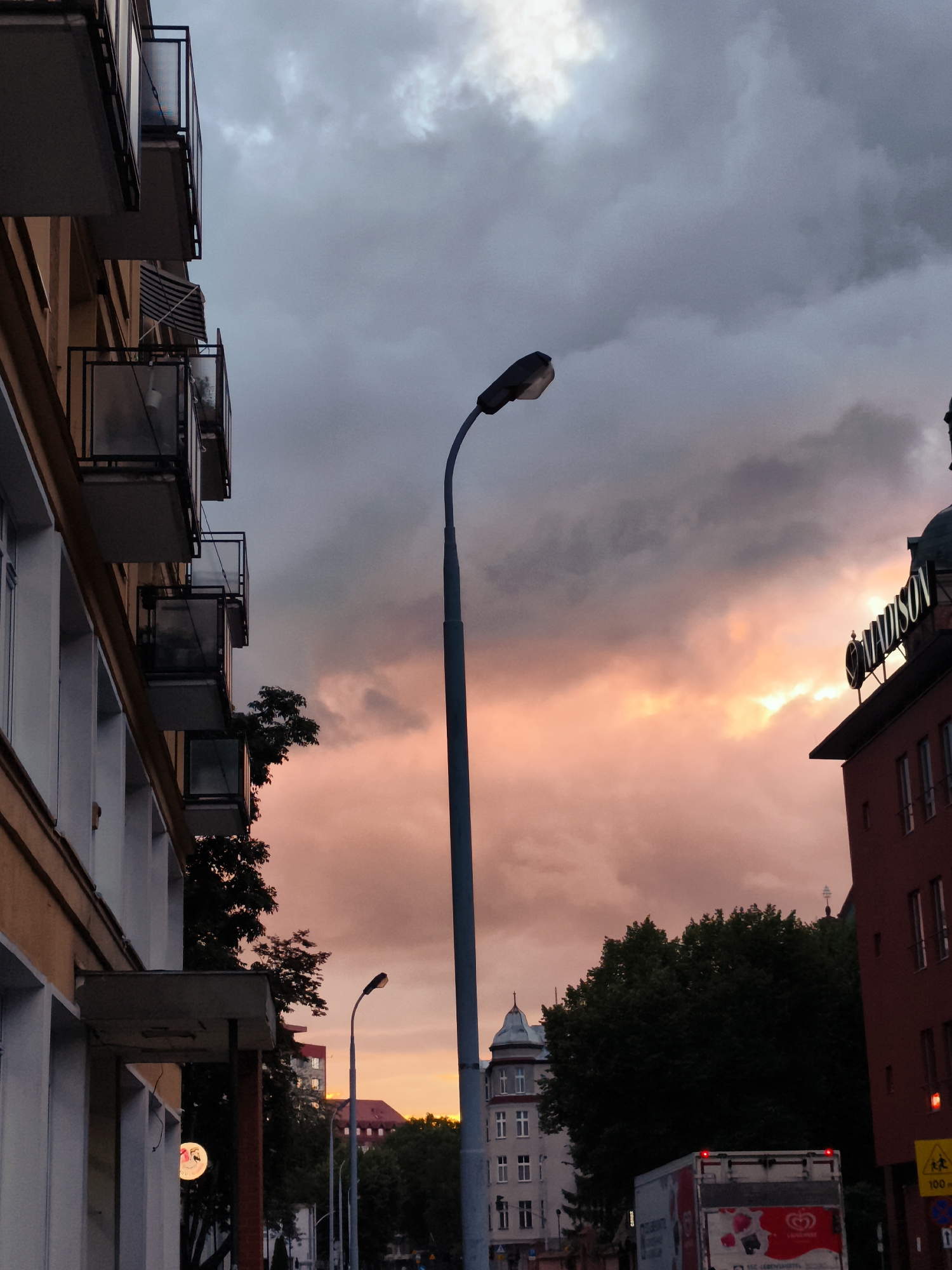 This image depicts a street scene in Gdańsk, Poland, during sunset. The sky is filled with dramatic clouds, transitioning from a dark, stormy gray at the top to hues of pink and orange near the horizon. A prominent street lamp stands tall in the foreground, its dark silhouette stark against the colorful sky. To the left, the side of a building with several balconies is visible, its architecture suggesting an older European style. On the right, a red building rises, topped with a sign that appears to read "MADISON." Further in the background, trees and other buildings can be seen, including a structure with a distinctive domed roof. A truck with its hazard lights illuminated is parked on the street, and there are traffic signs visible. The overall impression is a city street at dusk, with the vibrant colors of the sunset creating a striking contrast with the urban environment.