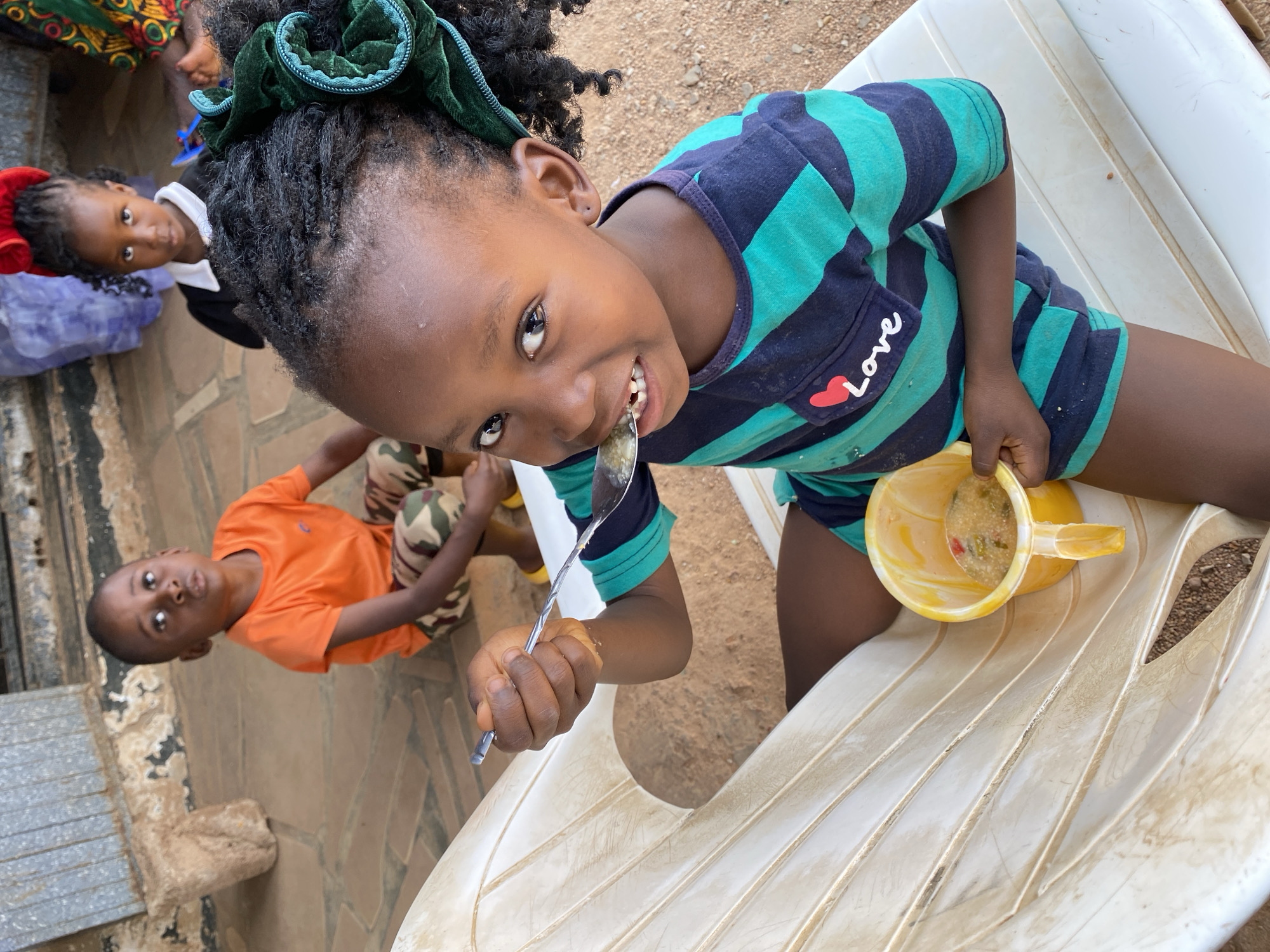 In Kuru Karama, Nigeria, a young girl sits on a white plastic chair outdoors. She wears a striped blue and green shirt with the word "LOVE" and a heart on the pocket, green hair tie, and has a spoonful of food raised to her mouth from a yellow plastic jug of stew. To the left, another child in an orange shirt and camouflage pants sits on the ground. Behind them, a third child with a red bow in their hair is partially visible. The ground is paved with stone.