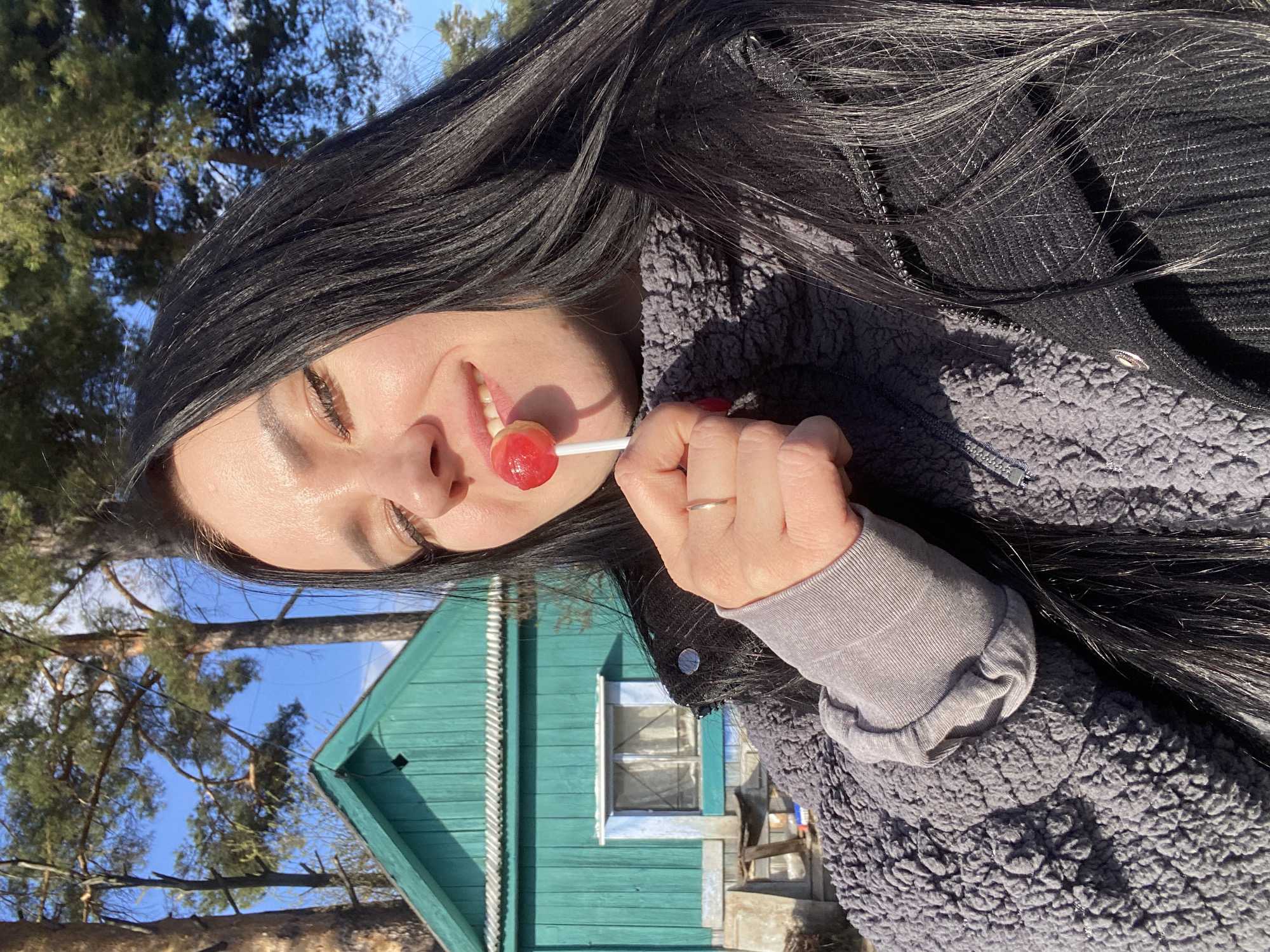 A woman with long dark hair is holding a red lollipop to her mouth. She is wearing a gray jacket and a gray long-sleeved shirt underneath. There is a light green wooden house with a white-framed window in the background. Trees and blue sky can be seen above the house. The location is in Chaikovsky, Russia.