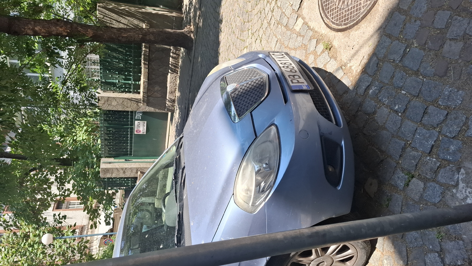 The image shows a blue car parked on a cobblestone street in Plovdiv, Bulgaria. The car is a Lancia Ypsilon, with a license plate reading "PB 6881KC". Behind the car, there is a green metal fence and a building. A street sign is visible on the fence, indicating "GARAGE NO PARKING". The scene is bathed in sunlight, casting shadows from trees onto the street.