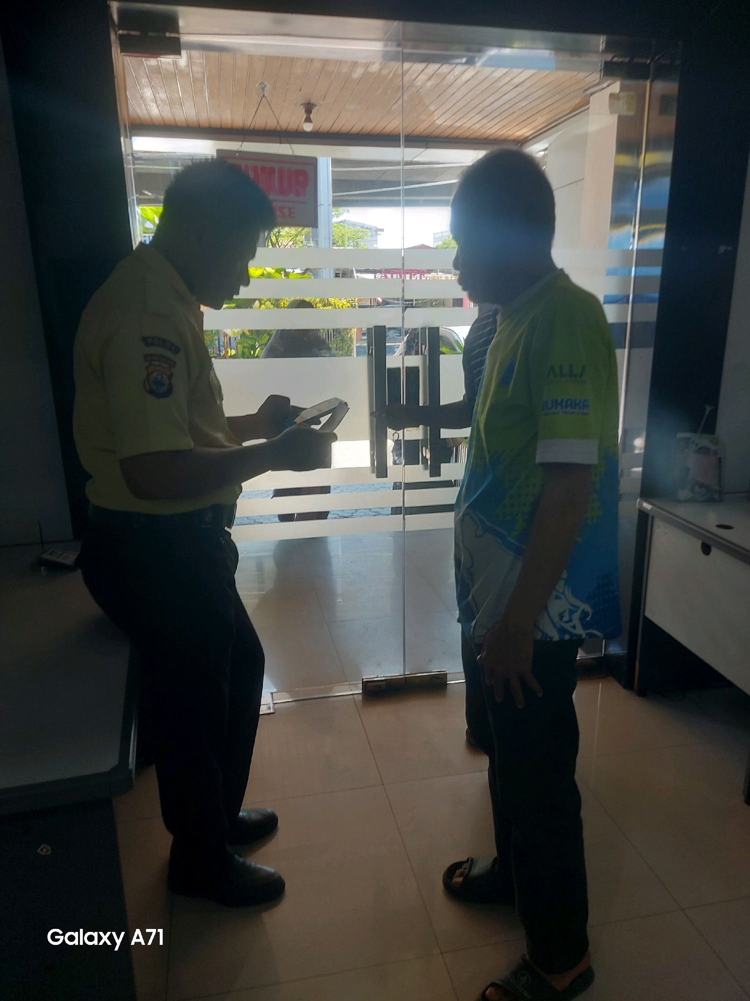 Two men are standing in front of a glass door, possibly an entrance to a building. The man on the left is wearing a light-colored uniform shirt with an emblem on the left sleeve and dark pants. He is holding a tablet or phone and appears to be looking at it. The man on the right is wearing a green and blue patterned t-shirt and dark pants with sandals. He is facing the man in the uniform and they appear to be interacting. Through the glass door, a sign with red lettering is visible, and the word "TUP" is partially discernible. The floor is tiled, and the lighting suggests it is daytime. The overall scene appears to be a public or commercial space.