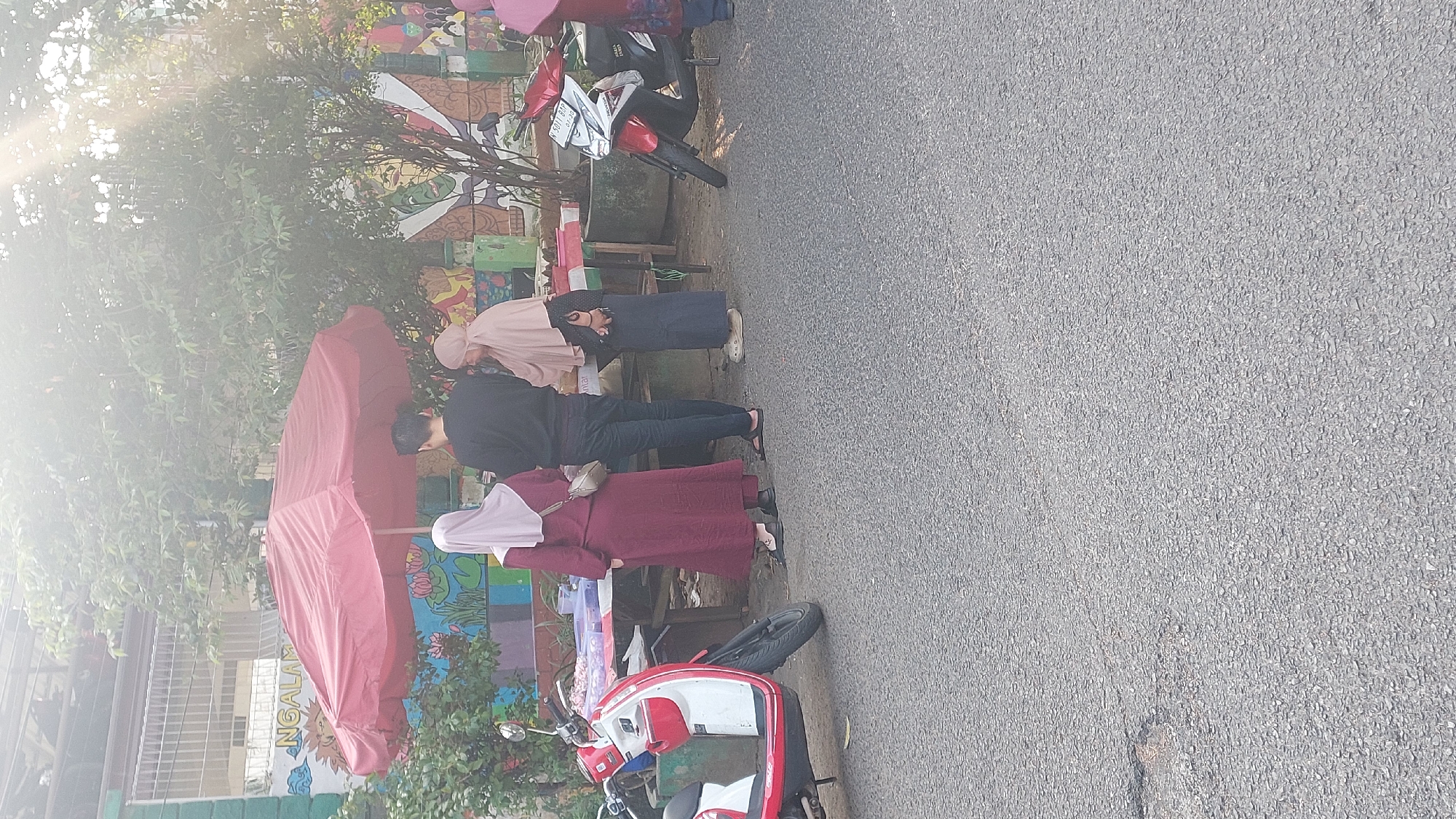 In Malang, Indonesia, a street scene unfolds under the bright morning sun. A vibrant red umbrella shelters a small roadside stall where two women and a man are gathered. The woman on the left, draped in a maroon hijab and dress, stands with her back to the camera. To her right, a man in a black long-sleeved shirt and dark trousers engages with the woman on the far right. This second woman, wearing a light brown hijab and a dark skirt, is focused on the items displayed on the stall, holding what appears to be a phone in her hand. The stall itself displays a variety of goods, possibly food items or small trinkets, laid out on a red and white striped cloth atop a makeshift table.
The background is a lively mural painted on a wall, depicting colorful flowers and other imagery, possibly part of a local art project or school. A white and red scooter is parked to the left of the stall, while a red and white motorcycle is positioned to the right, suggesting a common mode of transport in the area. The ground is paved with asphalt, showing signs of wear and tear with some cracks and unevenness. The overall impression is that of an everyday market scene in a bustling Indonesian city.
