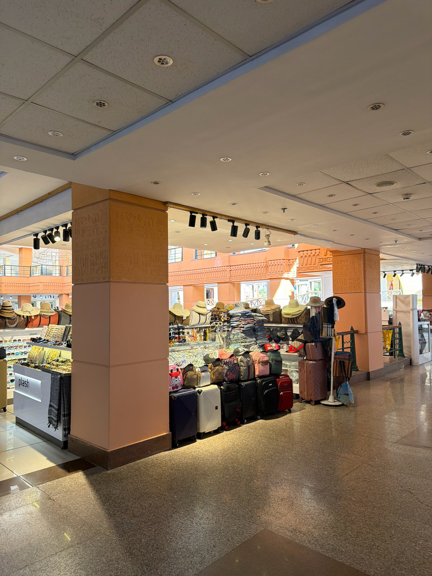 The image shows a retail shop interior, likely within a market or mall, in Sharm El Sheikh, Egypt. The shop specializes in accessories such as hats, sunglasses, and luggage. Prominently displayed are various styles of hats, including straw hats, some arranged on shelves and others on mannequins or stands. A display of sunglasses is also visible. Several suitcases and backpacks are stacked neatly, suggesting a focus on travel goods. The shop features a beige-colored pillar with hieroglyphic-like engravings, a design element often associated with Egyptian cultural themes, reinforcing the location. The ceiling is a standard drop ceiling with integrated lighting and ventilation grilles. The flooring is a polished, speckled tile. In the background, architectural elements with a similar beige, carved motif are visible, suggesting a consistent design theme throughout the establishment or the surrounding area. The overall impression is that of a bustling tourist-oriented retail space.
