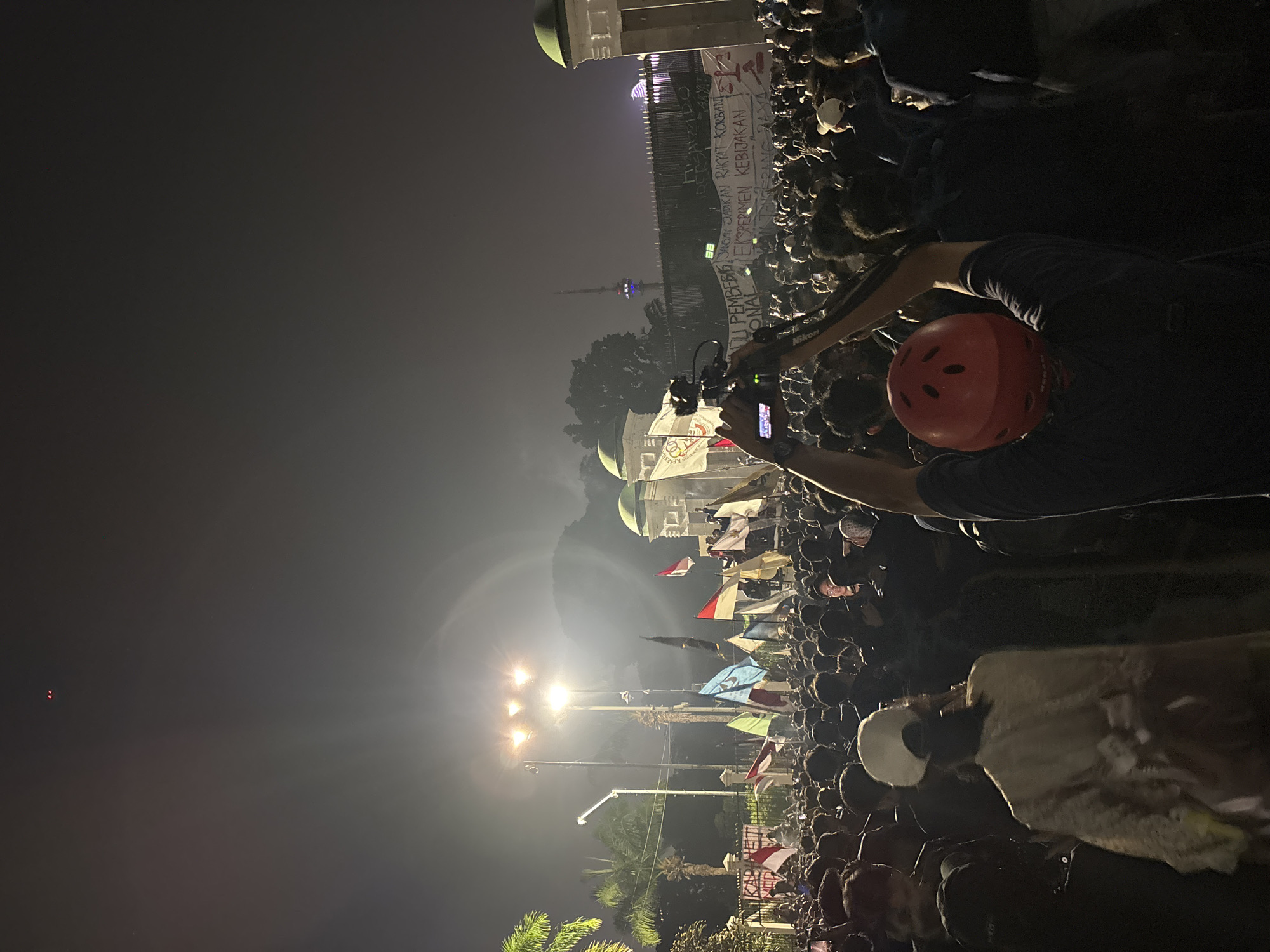 This media file depicts a nighttime gathering of a large crowd in what appears to be an urban setting in Central Jakarta, Indonesia. The scene is illuminated by artificial lights, including streetlights and lights from buildings, creating a somewhat hazy atmosphere.
The primary subjects are the densely packed crowd of people, many of whom are holding flags and banners, suggesting a protest, rally, or public event. In the foreground, a person wearing a red helmet is holding a camera and appears to be filming or photographing the event. Other individuals are also visible in the crowd, some with their faces obscured by darkness or distance.
In the background, a large building with green domes and intricate architectural details is prominent, possibly a mosque or a government building. A tall tower with colored lights is also visible in the distance. Several flags, predominantly red and white (the colors of the Indonesian flag), are being waved by the attendees. There is also a large banner stretched across the front of the building with text that is partially legible. Some of the visible text includes "WU PEMBE" and "RAKYAT KORBAN," which translates to "people the victim," and "EKSPERIMEN KEBIJAKAN NASIONAL," meaning "national policy experiment."
The time of day is clearly night, and the weather appears to be clear, though the lighting and atmospheric conditions create a sense of haze. The overall emotion conveyed is one of collective engagement and possibly a degree of intensity, given the large gathering and the presence of banners and flags. The location is strongly suggested to be Central Jakarta by the provided context.