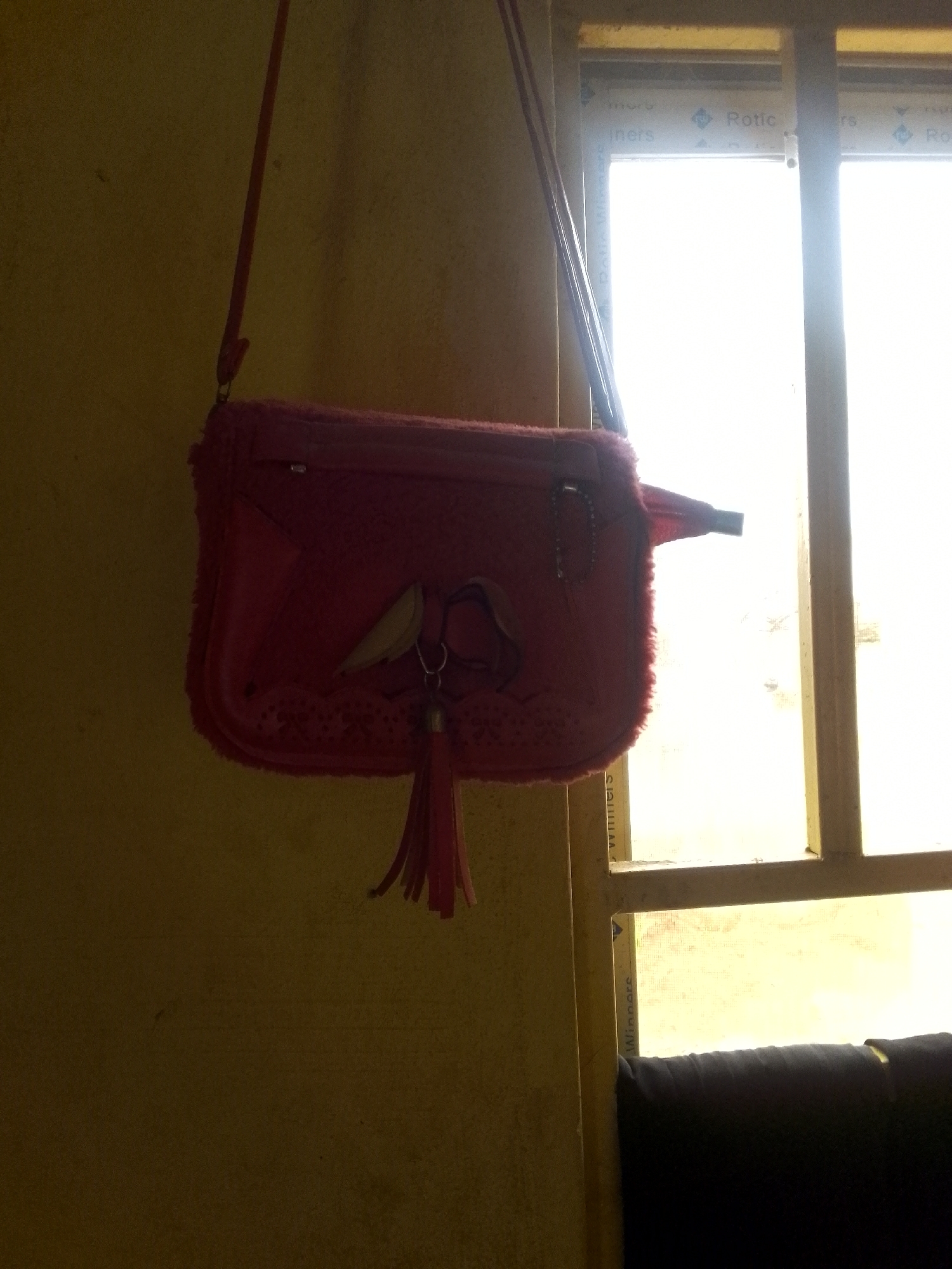 A close-up, eye-level shot captures a red handbag with fuzzy trim and a tassel hanging from the front, suspended against a yellow wall and a window with bright daylight shining through. The handbag has a decorative applique of what appears to be bunny ears. The window frame is a light yellow, and the glass panes are filled with a bright, blown-out white light, suggesting it is daytime. The wall to the left is a muted yellow with subtle texture. The scene is likely indoors, near a window. The image is slightly underexposed, especially on the left side, and the bright light from the window causes lens flare. There are no people visible, and the focus is on the handbag. The text "Rotico" and "Winners" are visible on a strip along the window pane.