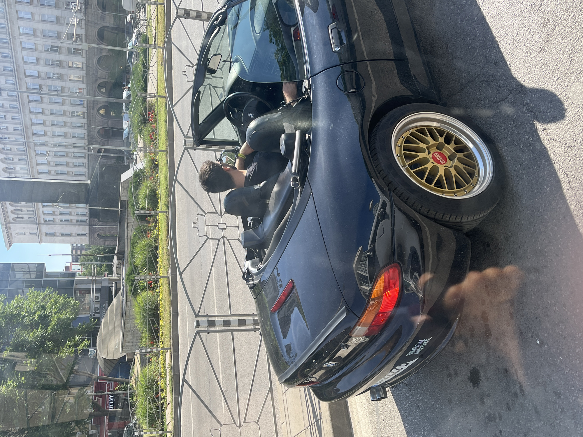 A man is driving a black convertible BMW Z3 on a city street. The car is facing away from the camera, and the man is wearing sunglasses and a black t-shirt. The car has a black convertible top and gold BBS rims. The street is paved with asphalt and there is a metal fence on the right side of the car. In the background, there are several buildings, including a large, ornate white building with many windows. The sky is blue and clear. There are trees and bushes along the side of the road. The overall impression is of a sunny day in a European city.