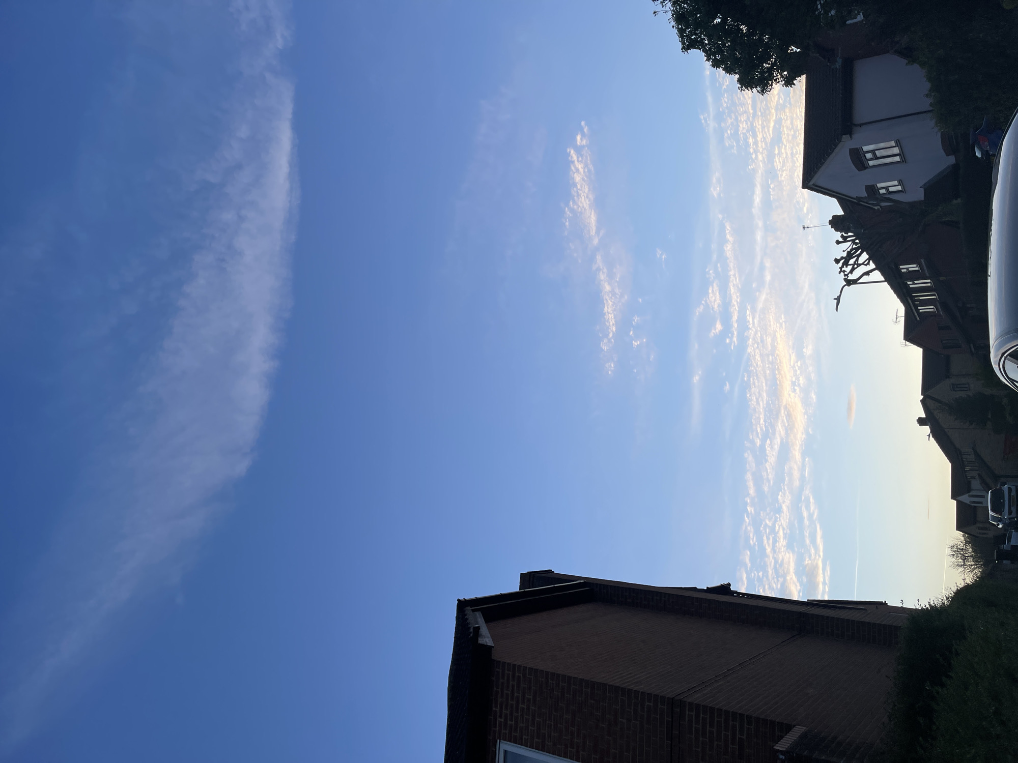 The image is a ground-level shot of a residential area in Maidstone, United Kingdom. The sky is predominantly blue, transitioning from a darker hue in the upper left to a lighter, sun-lit shade towards the upper right. Cirrus clouds are present, with a dense, streak-like formation on the left and more scattered, textured patterns on the right. The tops of brick buildings with slanted roofs and dormer windows are visible along the bottom and top edges of the frame. Green foliage, likely from trees and shrubs, appears in the upper right and bottom right corners. A few parked cars are seen in the upper right. The photograph is taken at an angle, making the scene appear tilted.