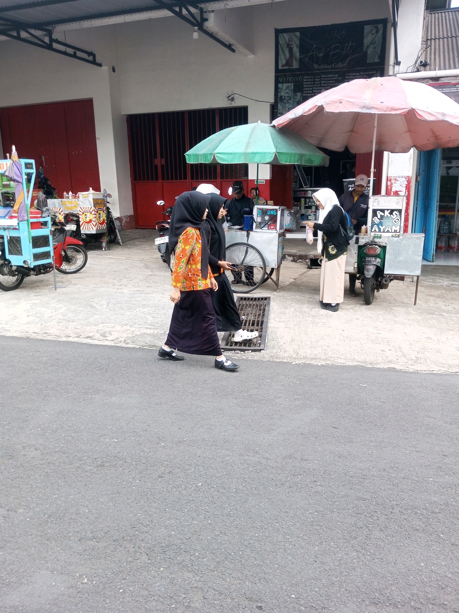 In the city of Krasak, Indonesia, a street scene unfolds with several individuals engaged in commerce and daily activities. A red motorcycle is parked, contributing to the urban landscape. In the foreground, a person walks on a paved road, while in the mid-ground, two individuals are in conversation near a food cart. Another person stands near a cart with a green umbrella, suggesting a market or roadside vendor setup. A stall displays various goods, and a motorcycle is parked nearby. The background reveals buildings with red doors and windows, characteristic of Indonesian architecture. The overall atmosphere depicts a bustling street in a local Indonesian community.