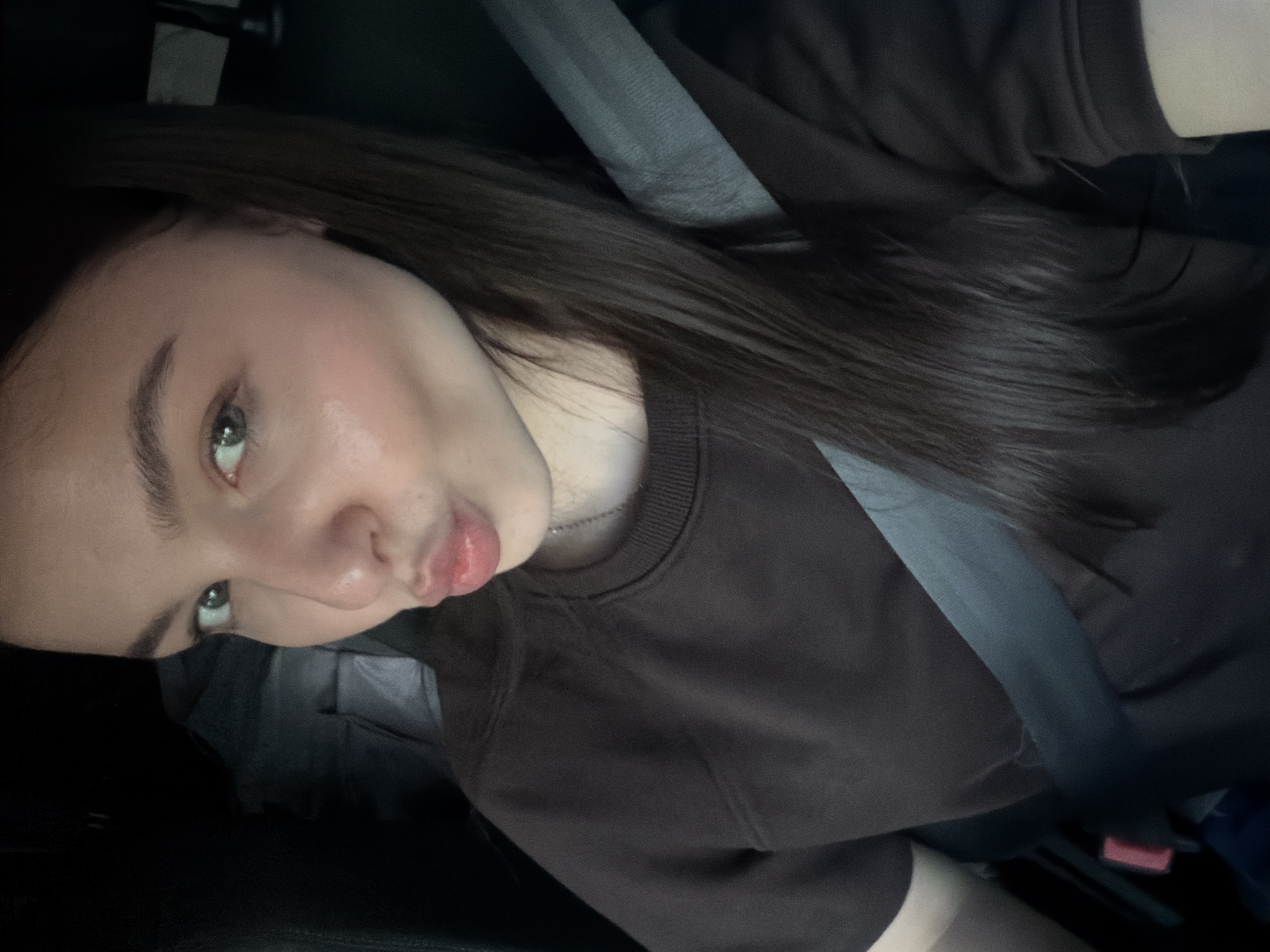 A female subject, appearing to be a young adult, is depicted in a close-up, chest-up portrait within what appears to be the interior of a vehicle. She possesses a fair complexion, long dark brown hair styled over her right shoulder, and light-colored irises (appearing hazel or green). Her lips are pursed forward, forming a "kiss" expression, and her gaze is directed towards the camera lens. She is wearing a dark brown, long-sleeved crew-neck top and a thin, silver-toned chain necklace. A grey automotive seatbelt is secured diagonally across her torso, extending from her right shoulder to her left hip, with the red release button of the buckle partially visible near her left hip. The background is uniformly dark and indistinct, consistent with the interior of a vehicle, featuring subtle dark upholstery textures. The specific location in Sofia, Bulgaria, cannot be determined from the visual information provided within the image.