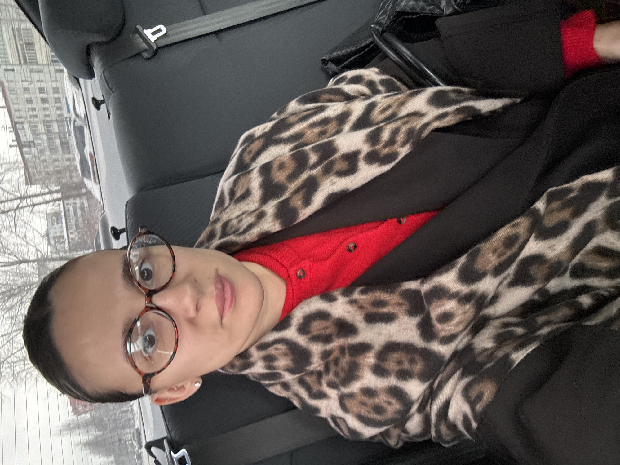 A person with round glasses is in the back seat of a car, wearing a red sweater, black jacket, and a leopard-print scarf. Buildings are visible through the window. Based on the location request, it is likely that the location is Chaikovsky, Russia.