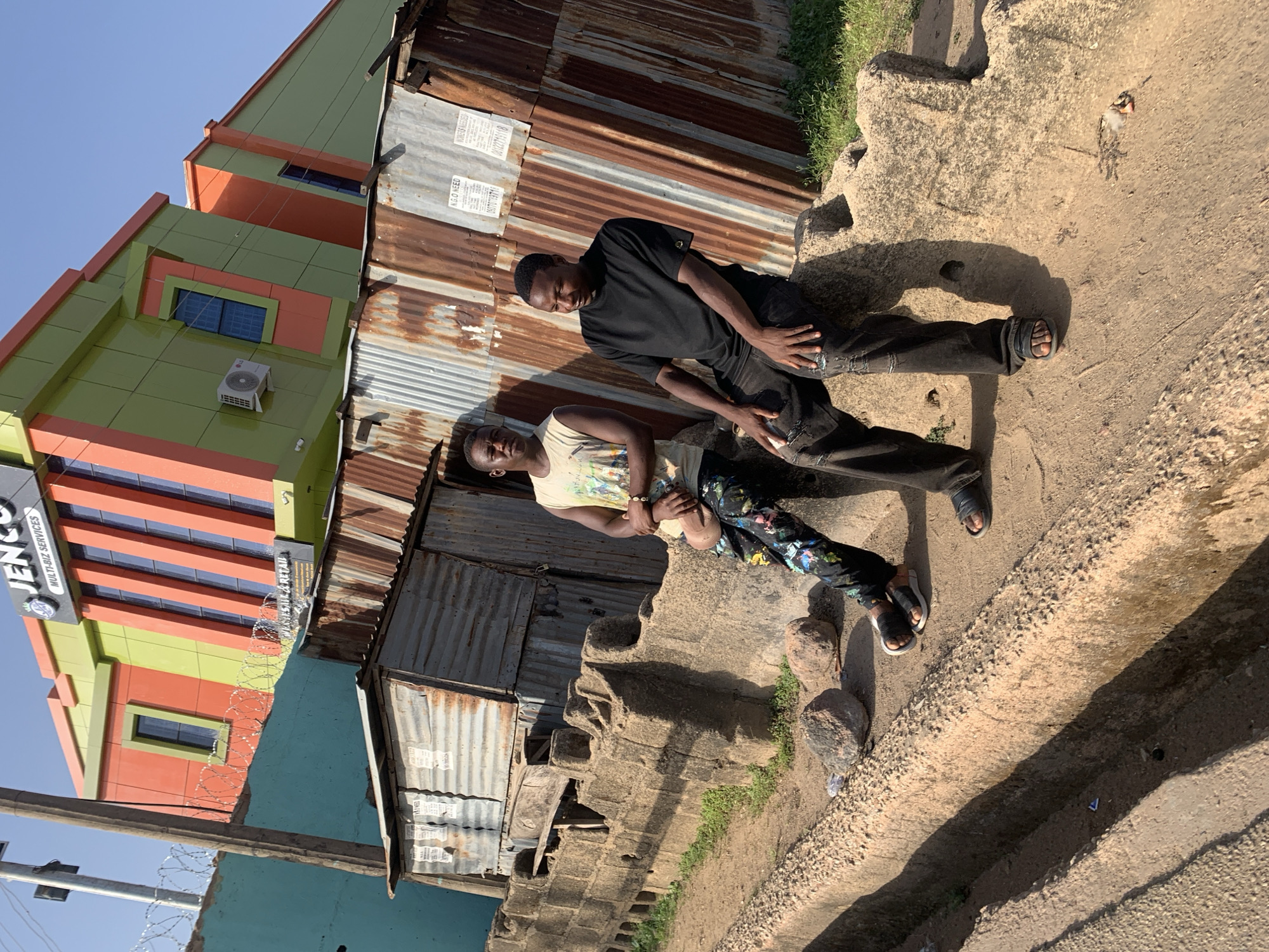 The image shows two young men posing in front of a corrugated iron building. One man is wearing a black shirt and dark pants, and the other is wearing a light-colored t-shirt and patterned pants. They are standing on a dirt and concrete surface. To the left, a multi-story building with orange and green facade is visible, and a sign reads "JENCO Multi-Biz Services." The setting appears to be urban, possibly Jos, Nigeria. Barbed wire runs along the side of the corrugated iron building, and there are rocks and sparse vegetation on the ground. The light suggests it is daytime.