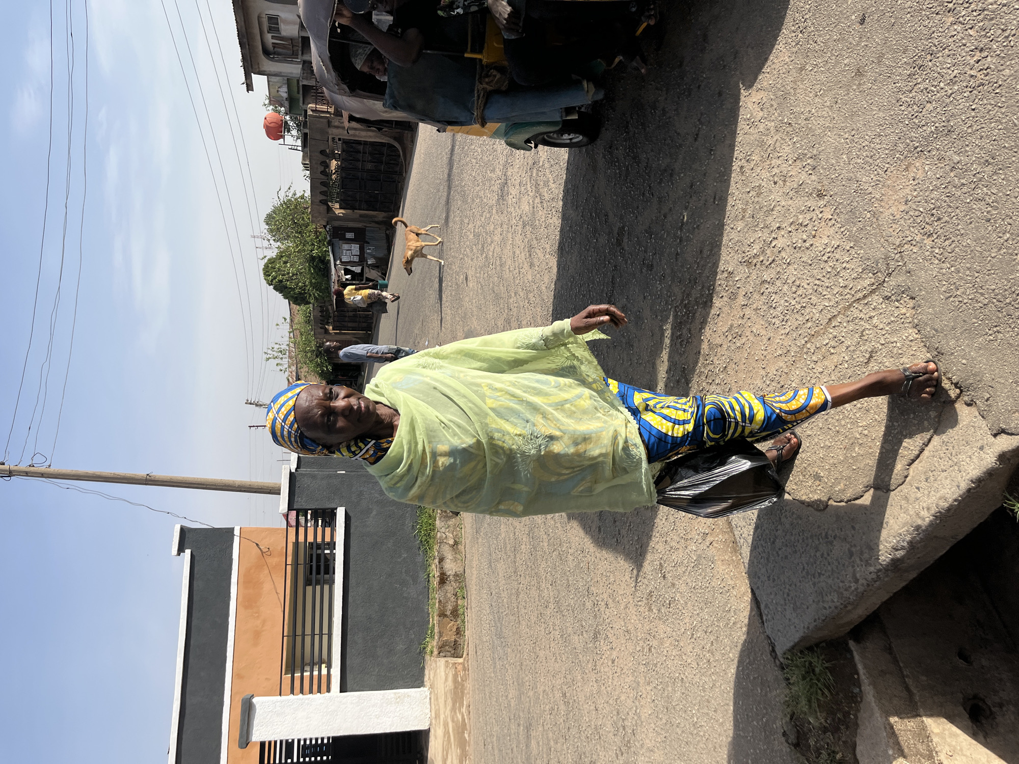 An elderly woman stands on the side of a paved road in Jos, Nigeria. She is wearing a headscarf, a light green shawl, and brightly colored patterned clothing. The location appears to be a residential street with a building on the left. In the background, a dog crosses the road and a vehicle is parked. The woman's arm is outstretched, and she is standing next to a concrete drainage ditch. The image suggests an outdoor scene during daylight.