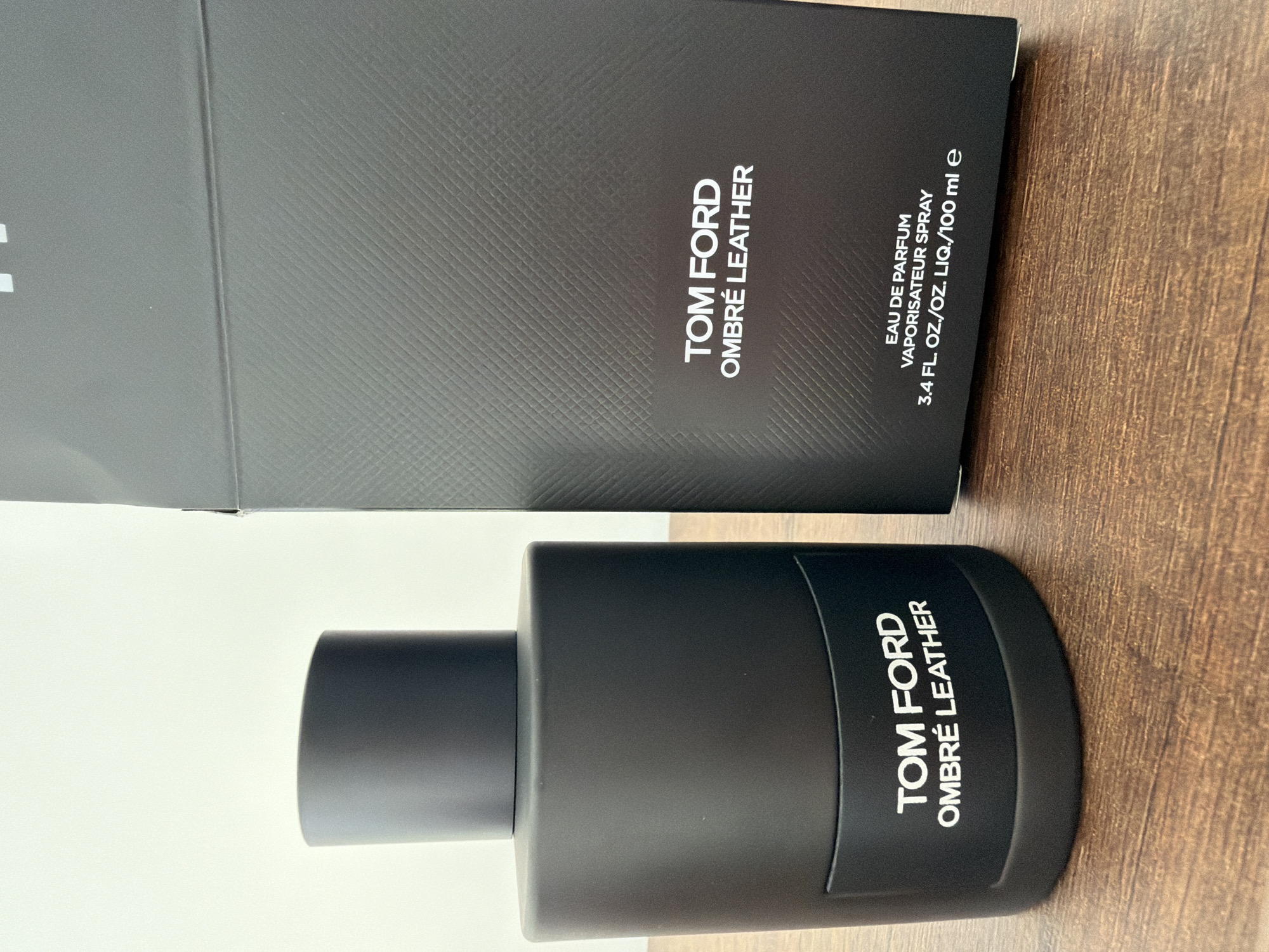 A black Tom Ford Ombré Leather fragrance bottle and its black textured box are displayed on a wooden surface. The bottle is cylindrical with a matte black finish and a black cap. White text on the bottle reads "TOM FORD OMBRÉ LEATHER". The box is placed to the right of the bottle and features the same textured pattern. White text on the box reads "TOM FORD OMBRÉ LEATHER" and further down, "EAU DE PARFUM VAPORISATEUR SPRAY 3.4 FL. OZ./OZ. LIQ./100 ml e". The background is a plain white wall. There are no people or animals visible in the image, and no indications of a specific location in Finestrat, Spain.