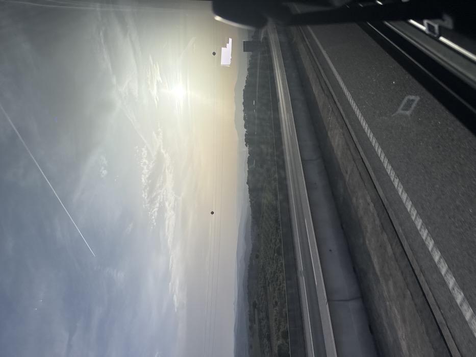 The image depicts a panoramic view of an elevated highway cutting through a rural landscape, observed from the interior of a vehicle, likely in Macedo de Cavaleiros, Portugal. The scene is illuminated by a low sun, positioned in the mid-right, indicating either sunrise or sunset. The sky, a hazy pale blue to yellow gradient, features scattered cirrus clouds and a prominent white contrail stretching from the upper left towards the upper right.
In the mid-ground, a multi-lane asphalt highway is visible, separated by a concrete median barrier. White lane markings, both solid and dashed, are present on the road surface. Below the highway's elevation, the landscape consists of rolling hills covered with dense, dark green vegetation, possibly agricultural land or scrub, extending into a misty horizon.
Several high-voltage power lines with spherical bird diverters traverse horizontally across the mid-ground above the landscape. One distinct dark spherical marker is visible near the center-left. On the far right, a dark, vertical structure, possibly part of a building or tower, stands against the horizon. The immediate foreground shows a dark vehicle interior component, likely a headrest or pillar, on the right. Reflections and lens flare from the sun are evident on the viewing surface, including a faint diamond shape reflected on the asphalt in the lower right. No individuals or specific actions are discernible within the frame.