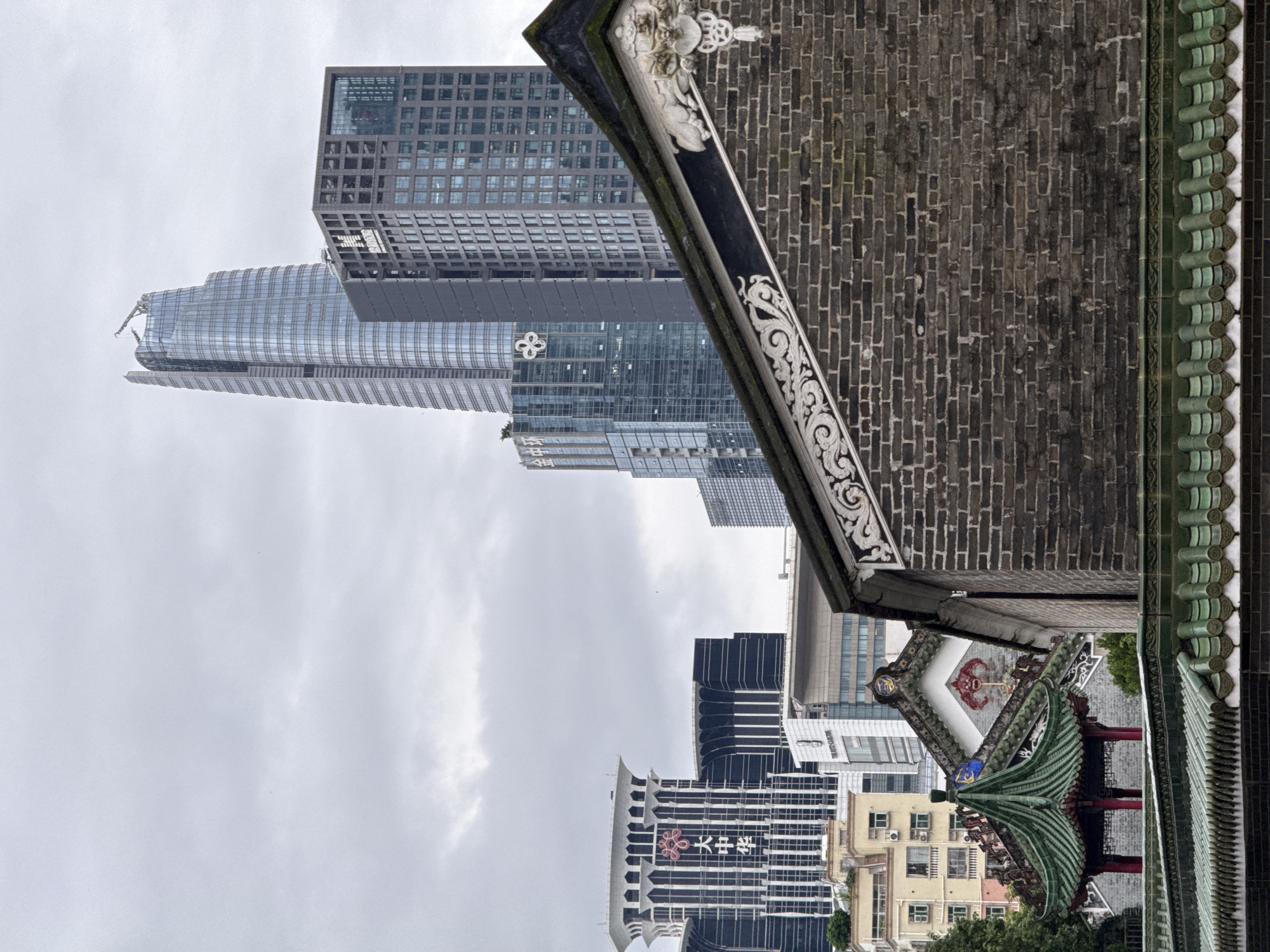 The image captures a view in Futian District, China, featuring a juxtaposition of traditional Chinese architecture and modern skyscrapers. In the foreground, the dark brick facade of a traditional building is visible, adorned with intricate white decorative carvings along its angled roofline. Below this, a row of green, glazed tiles forms the edge of another roof. Further back, a colorful, multi-tiered pavilion with a green, curved roof and red pillars stands out. Behind these traditional structures, a cluster of contemporary high-rise buildings dominates the skyline. The tallest among them is a sleek, glass-encased skyscraper with a distinctive, tapering top. To its right, a squarer, darker skyscraper features a grid-like pattern of windows. Other modern buildings of varying heights and architectural styles are also visible, some with illuminated signage. The sky is overcast with a pale, diffused light. There are no people or animals visible in the frame.