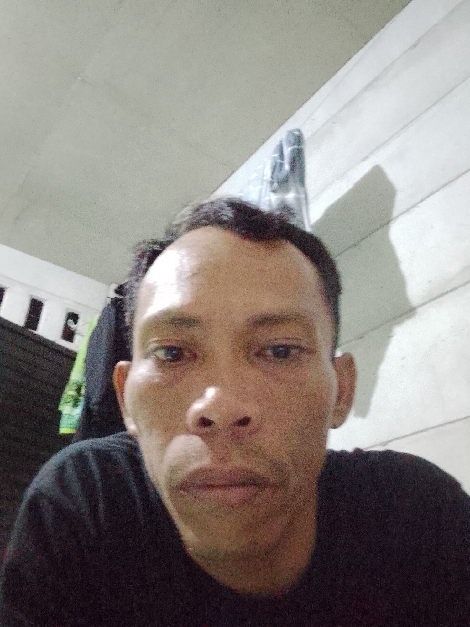 A man is centered in the frame, wearing a black t-shirt. The background includes a white ceiling and a paneled wall. Hanging items, including clothing and a towel, are partially visible on the left. The image appears to be taken indoors, possibly in a residential setting, Bukit Ketok, Indonesia.